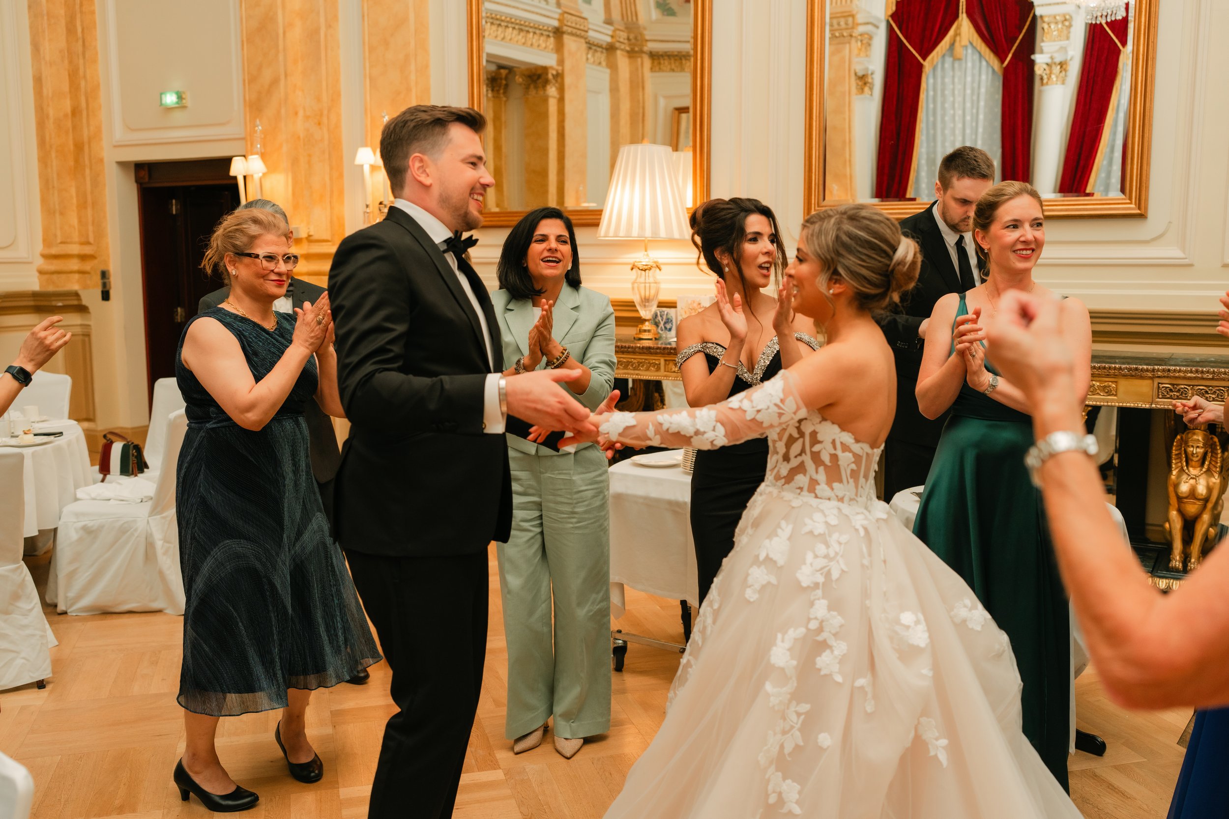 People celebrating at a formal event, with a bride in a white gown dancing with a man in a suit, surrounded by smiling and clapping guests in elegant attire.