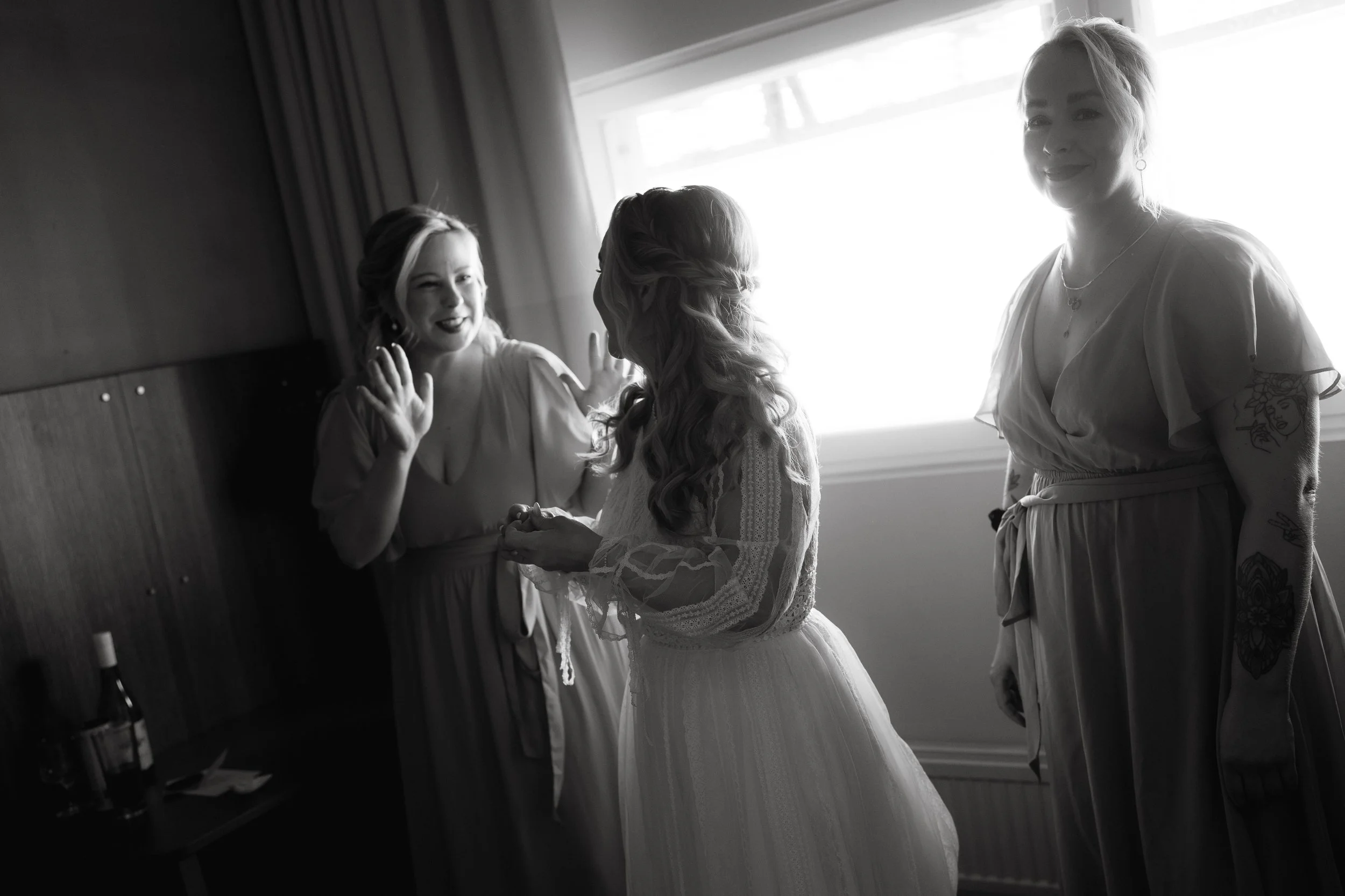 Black and white photo of a bride with two bridesmaids in a room, one with tattoos visible, standing near a window, all wearing dresses.