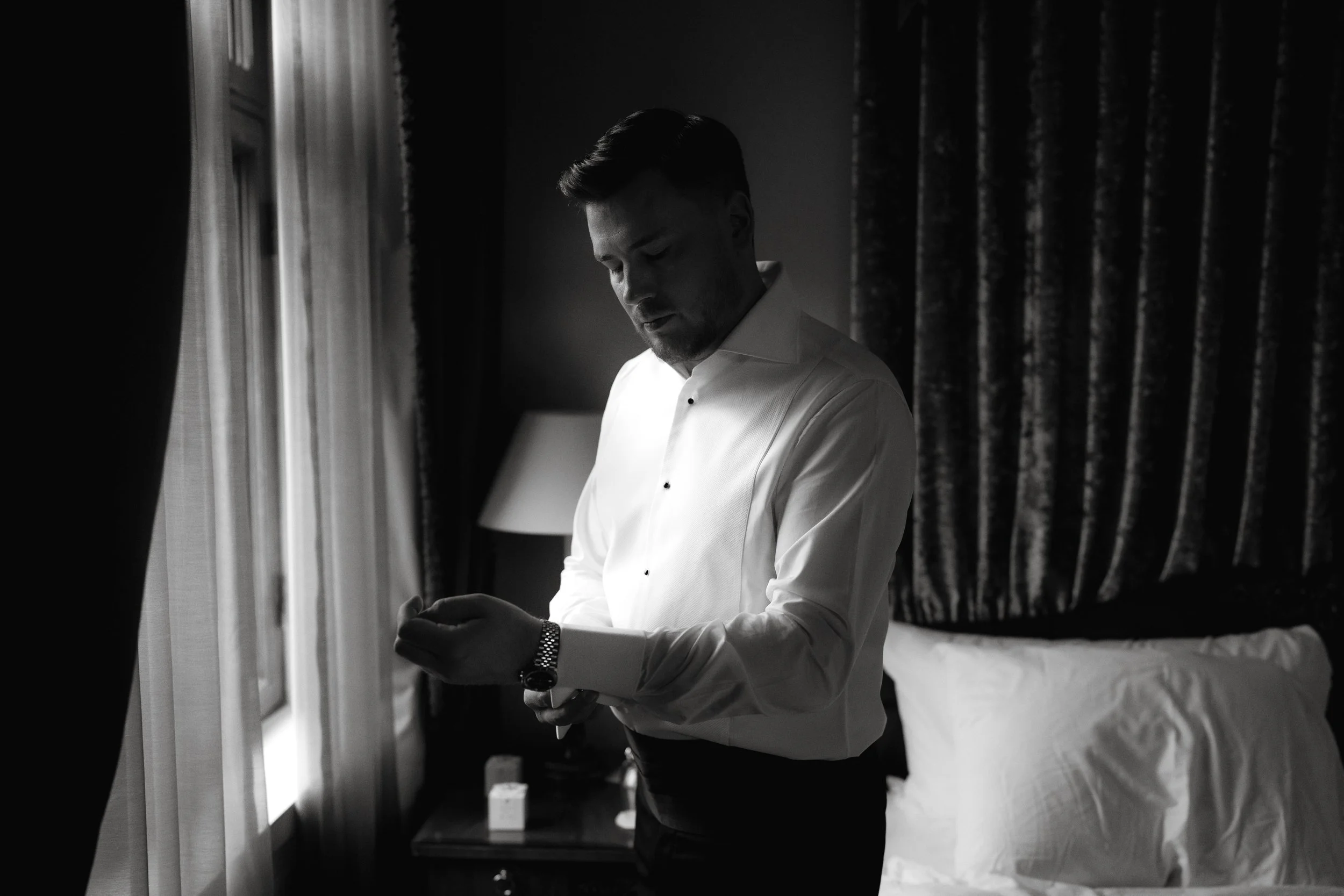 Man adjusting cufflinks, wearing a formal white shirt in a dimly lit room with curtains and bed.