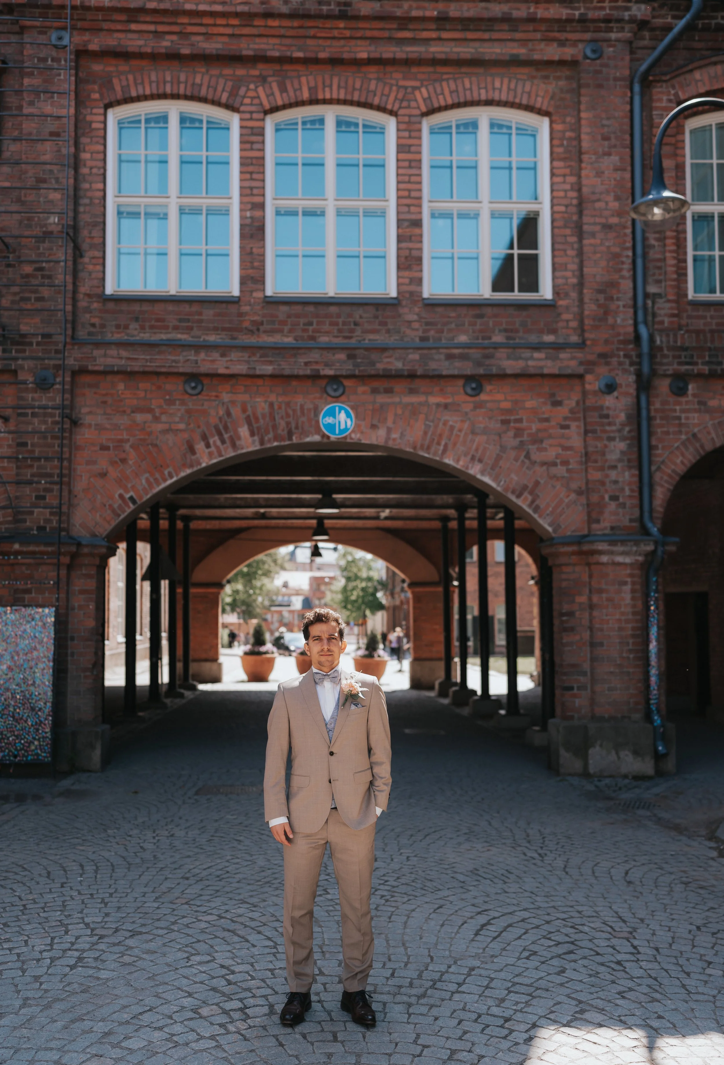 Man in tan suit standing on cobblestone under brick archway.