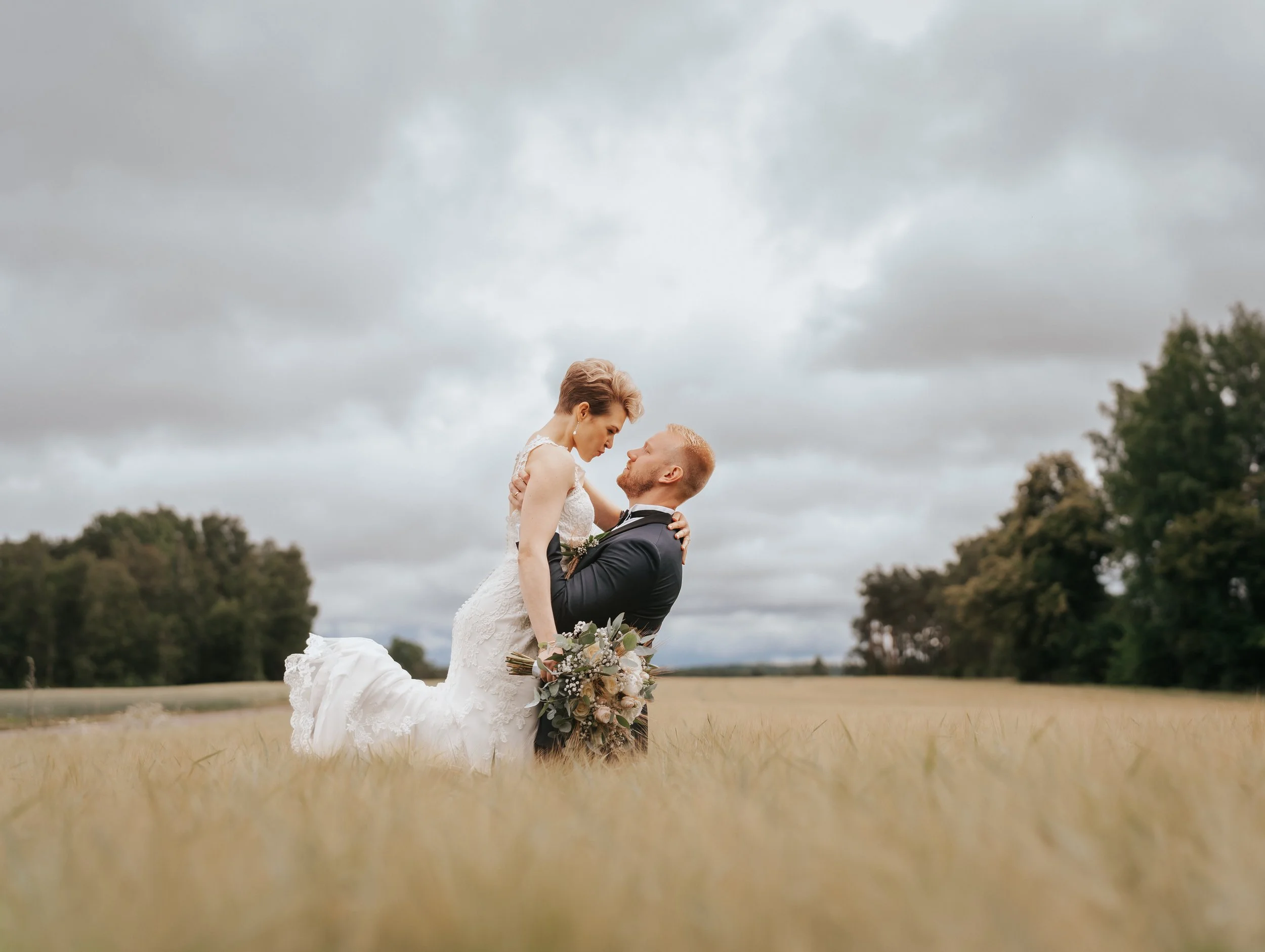A bride and groom embracing in a field with a cloudy sky.