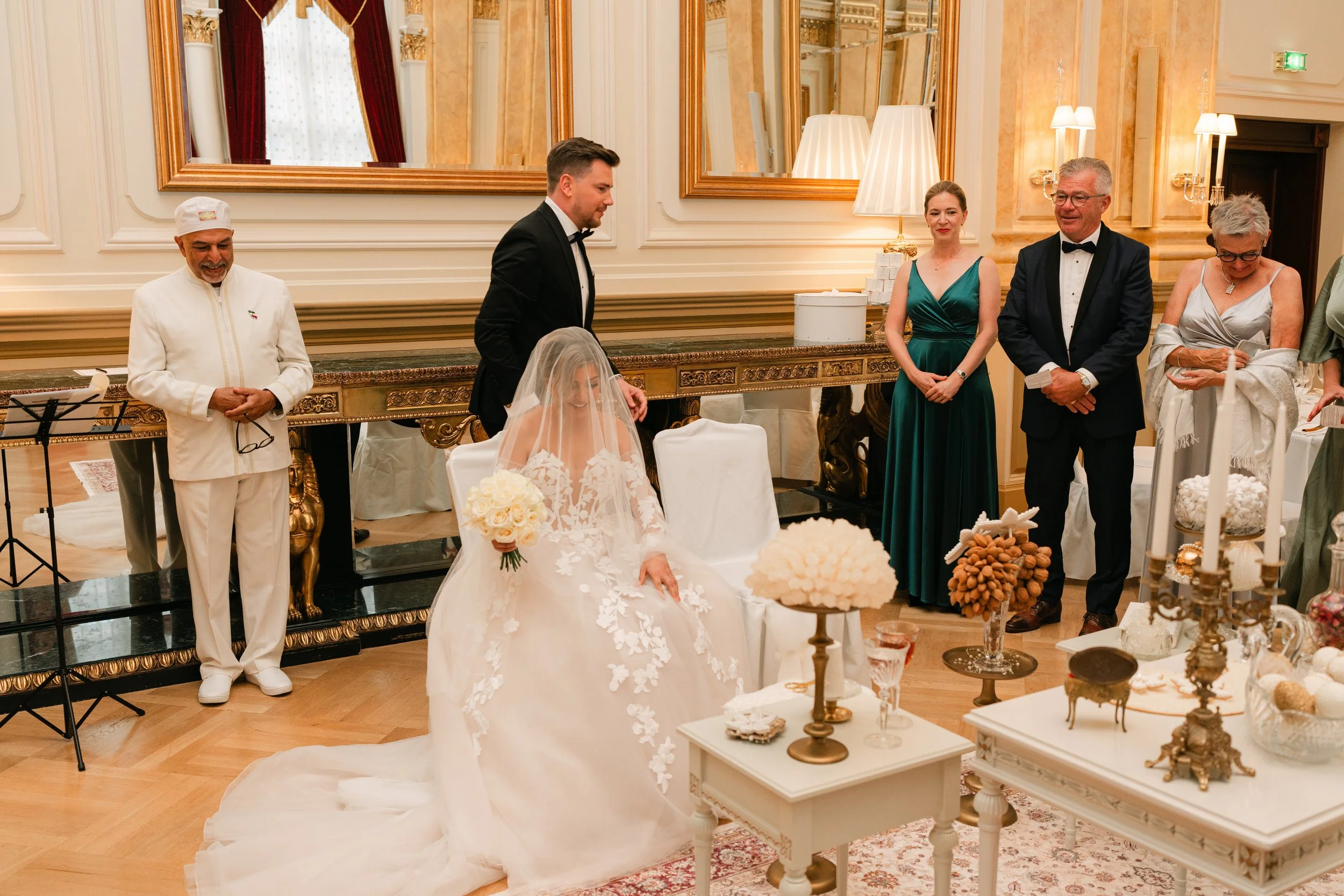 Elegant indoor wedding ceremony with bride in white gown and veil, groom in black tuxedo, and guests in formal attire, in a lavishly decorated room with ornate mirrors and furniture.