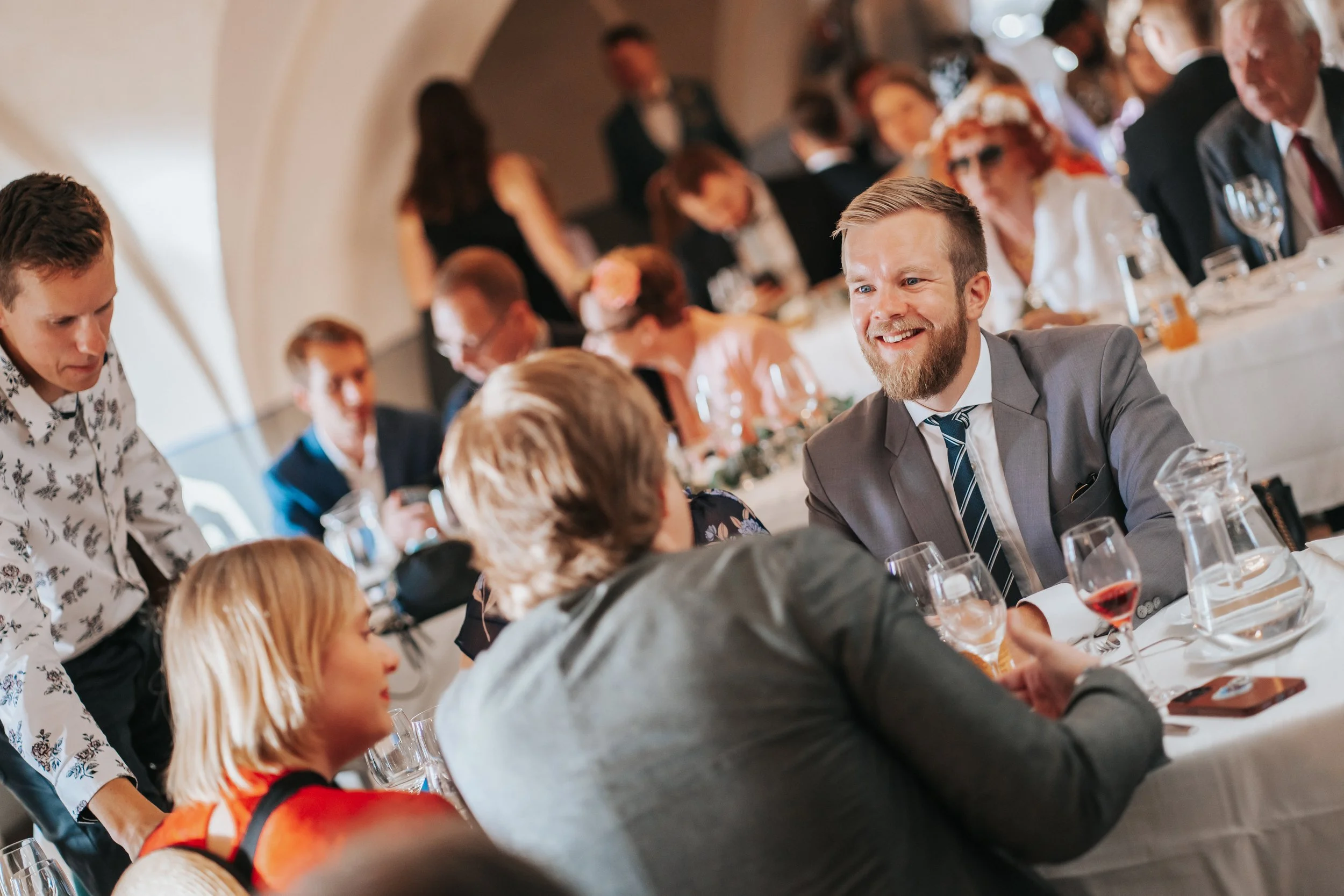 Group of people at a formal dining event, dressed in suits and dresses, seated at tables, with wine glasses and tableware.
