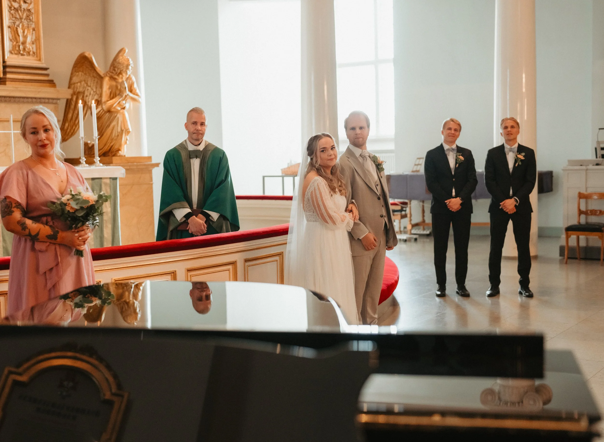 Bride and groom standing together with wedding party and priest in a church.