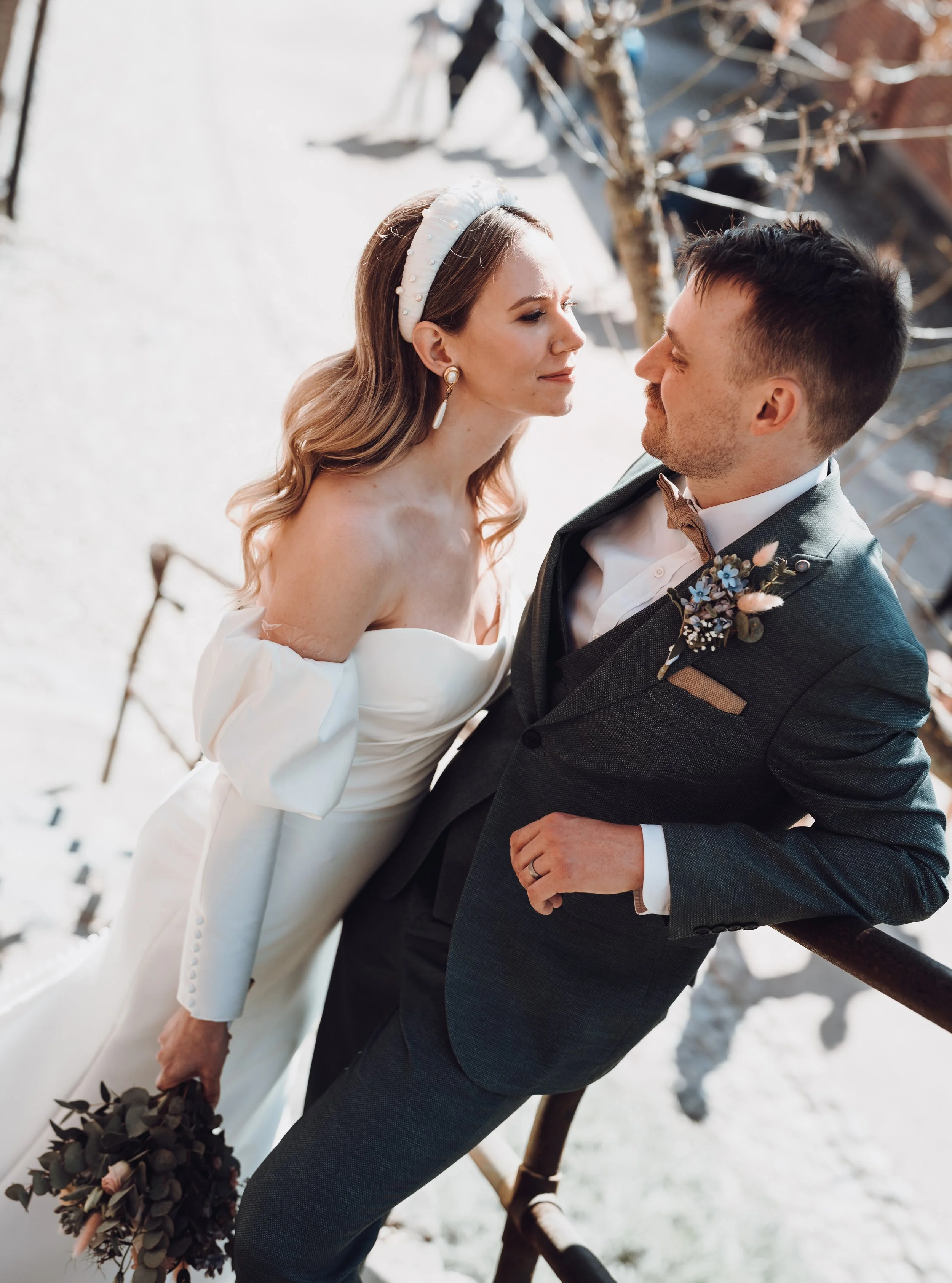 Bride in white gown with bouquet, groom in dark suit with boutonnière, smiling at each other outdoors.