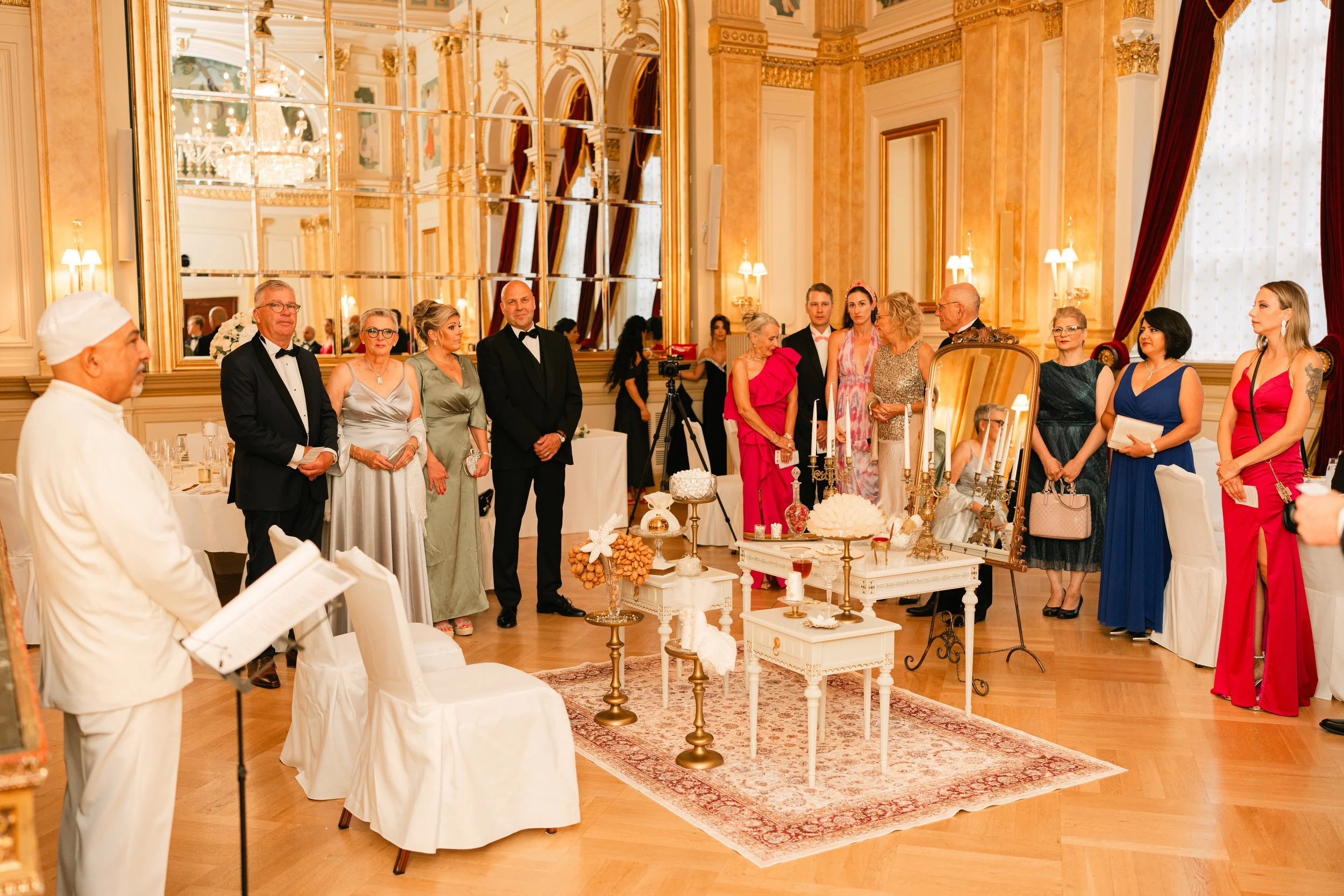 Elegant gathering in a luxurious room with people dressed formally, intricate decor, mirrors, and chandeliers.