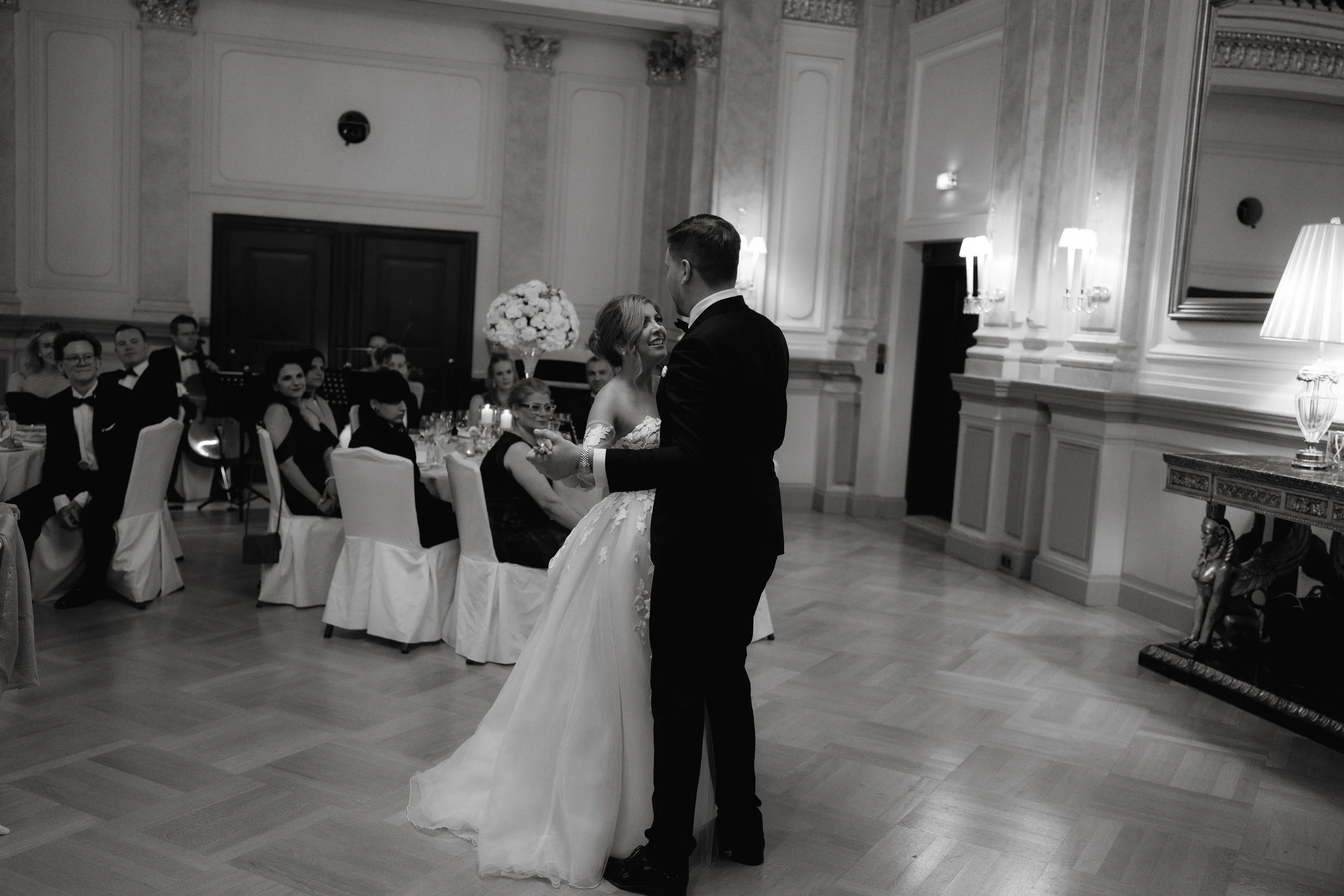 Bride and groom dancing at wedding reception in elegant ballroom, guests seated at tables, black and white photo.