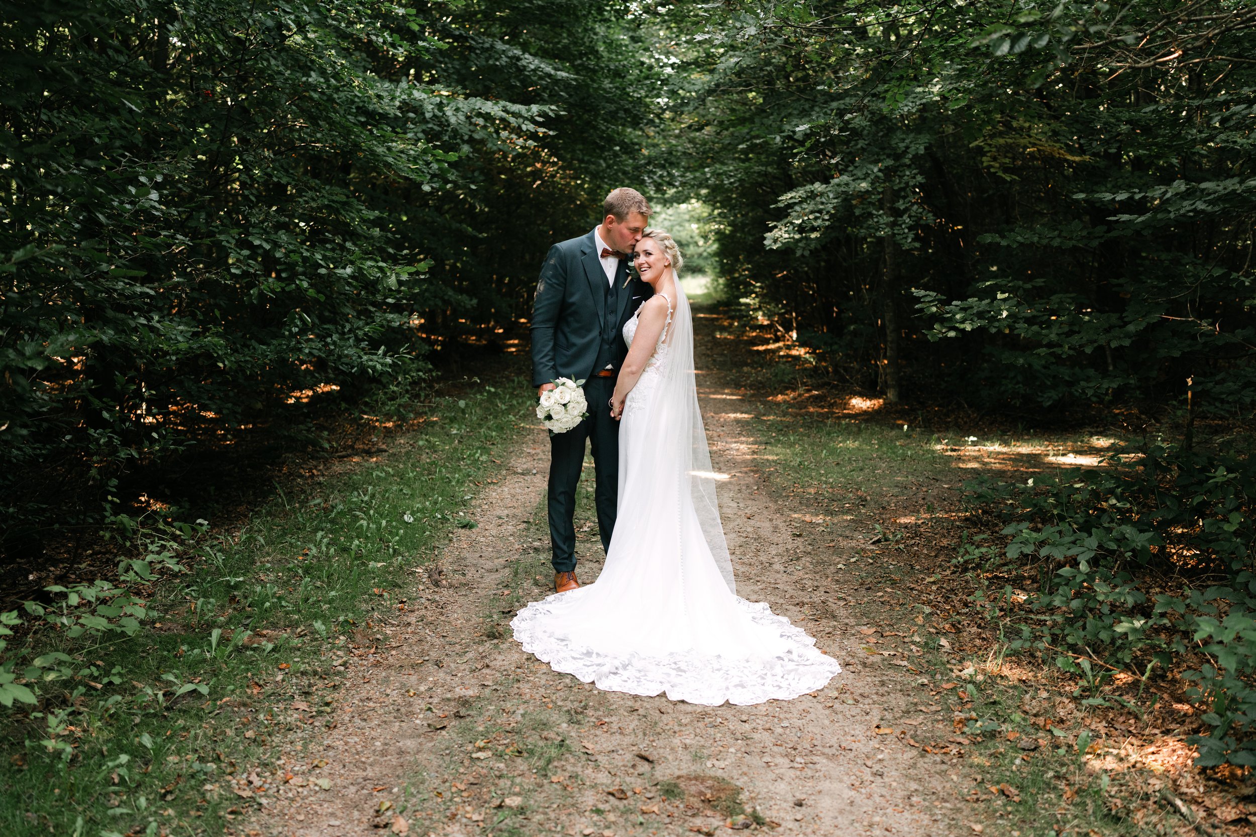 Bride and groom posing on a forest path, bride in white dress holding bouquet, groom in suit.
