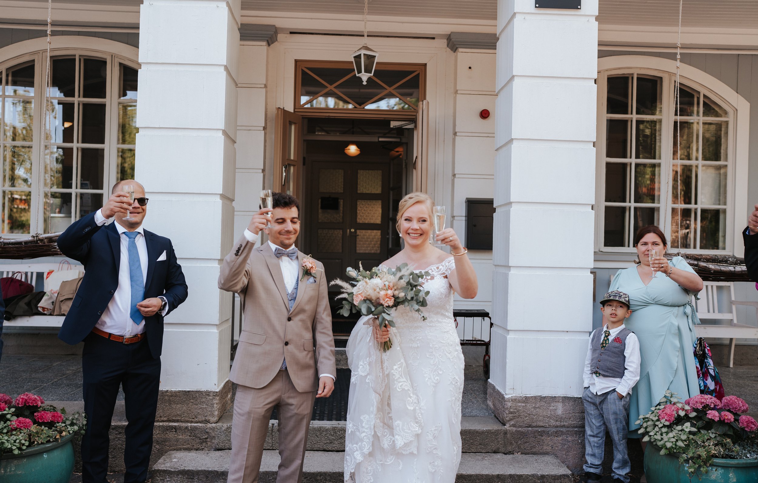 Bride and groom with guests holding glasses for a toast, standing outside a building.