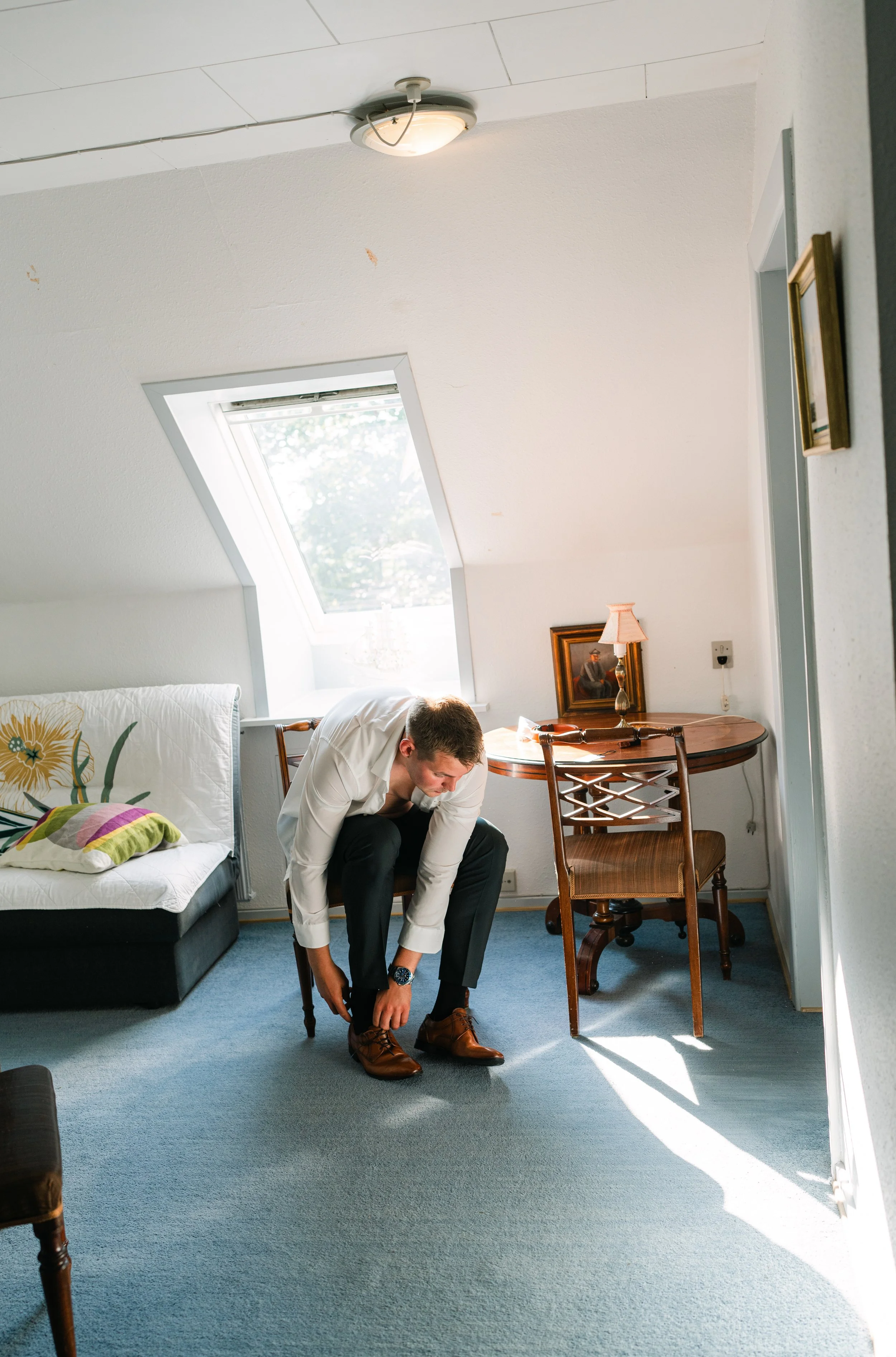 Man tying shoelaces in a sunlit room with a slanted ceiling, sofa, and table.