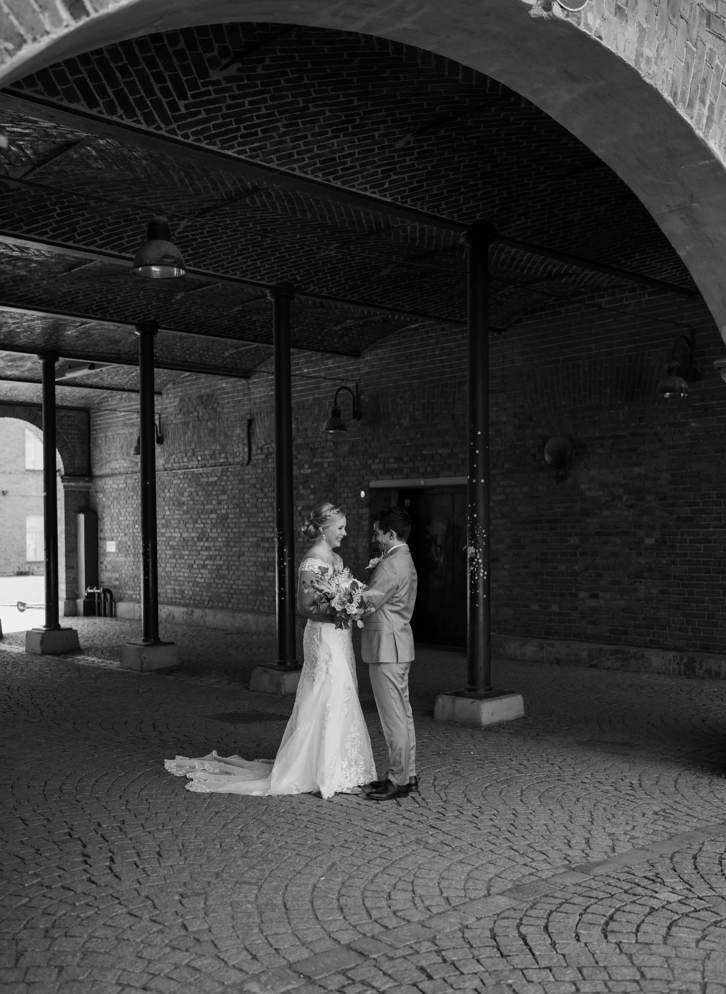 Bride and groom standing in a brick archway, holding hands and exchanging flowers, in a black and white photo.
