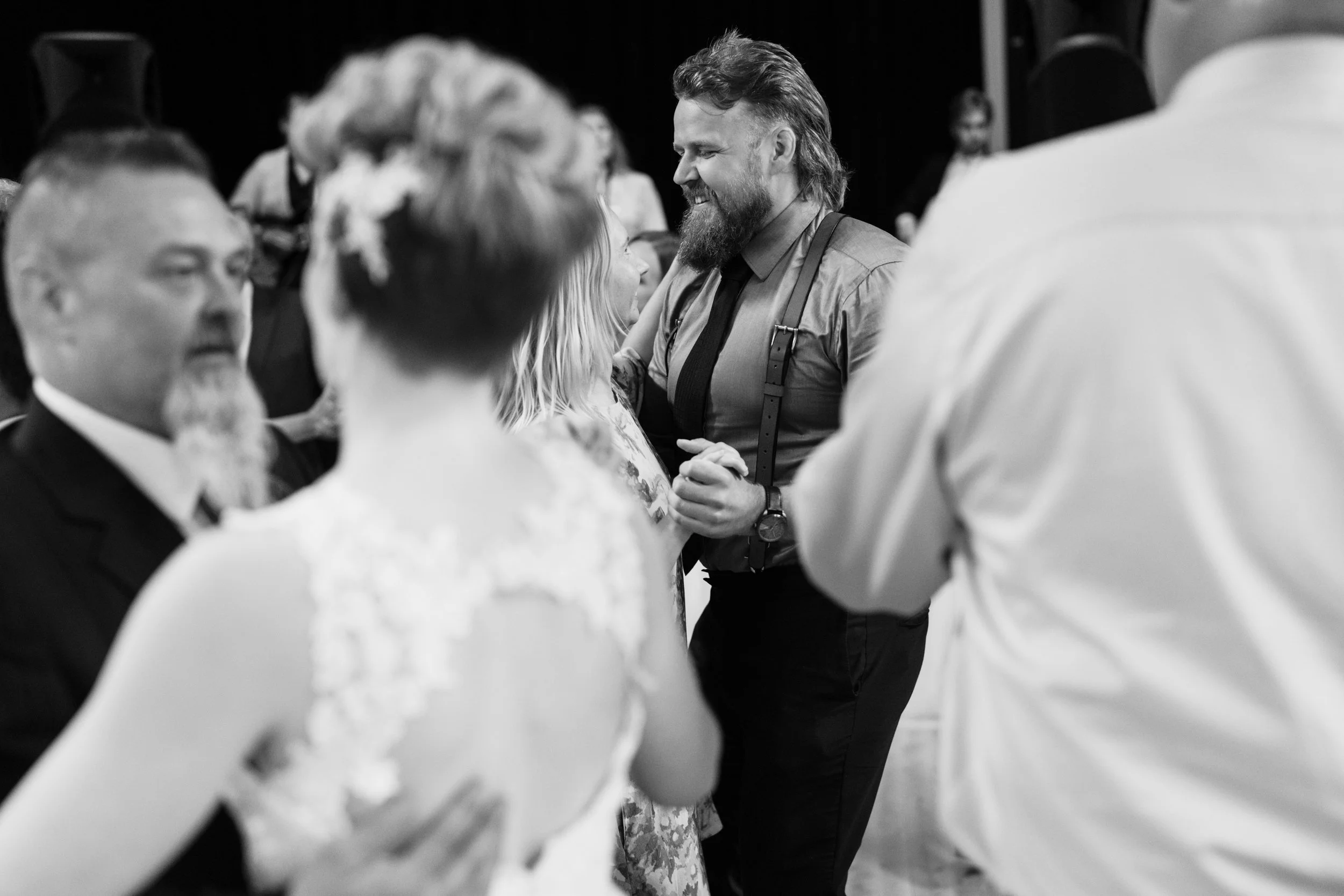 Black and white photo of people dancing at an event, including a bearded man in suspenders smiling while dancing.