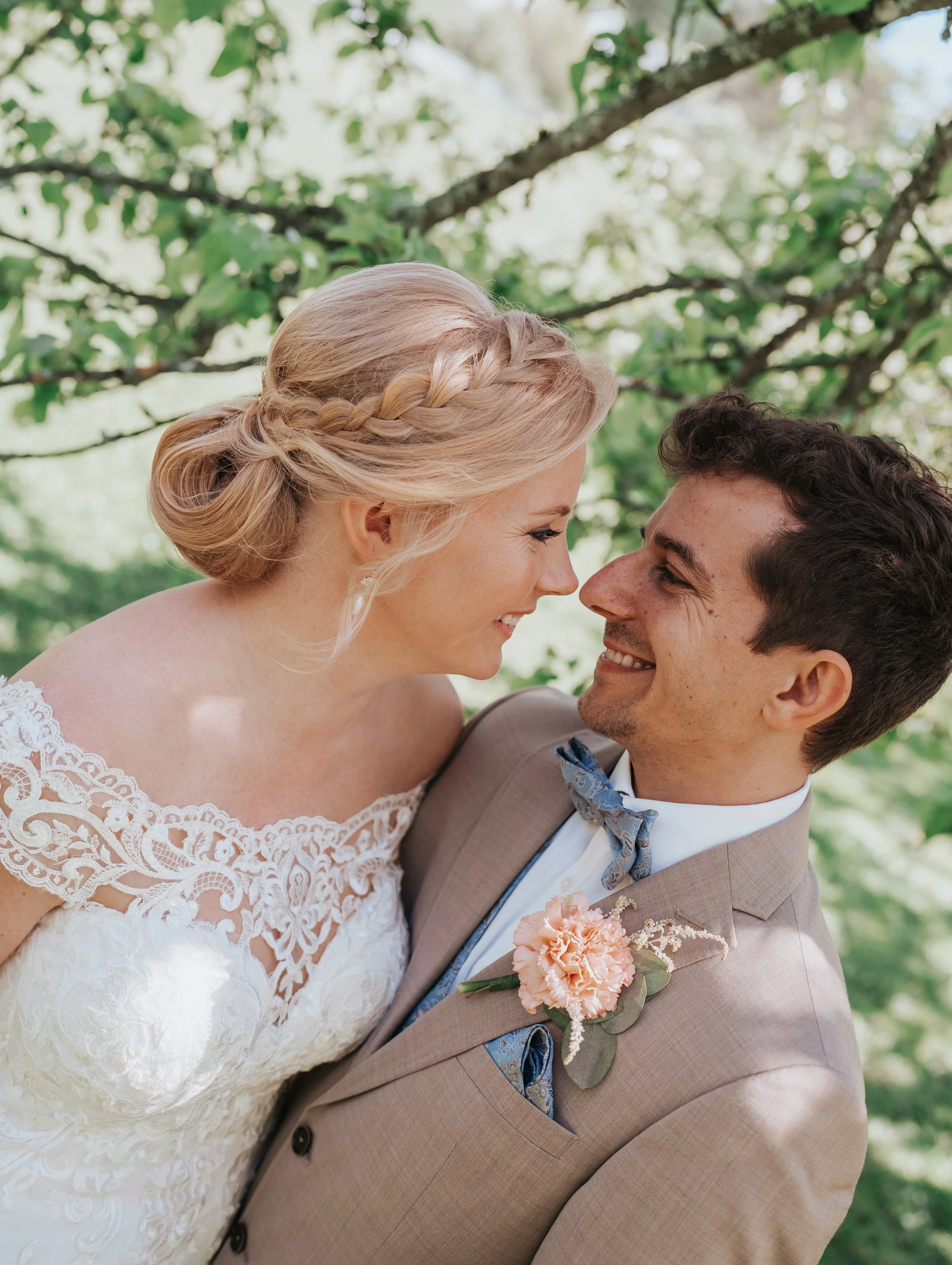 Bride and groom smiling at each other in a garden setting, bride wearing lace gown, groom in light suit with floral boutonniere.