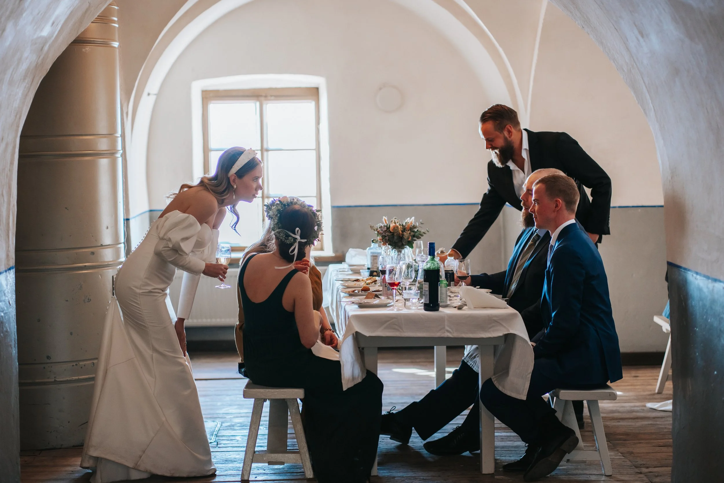 Wedding reception group sitting at a table in a historical building with a bride and groom talking to guests over drinks.