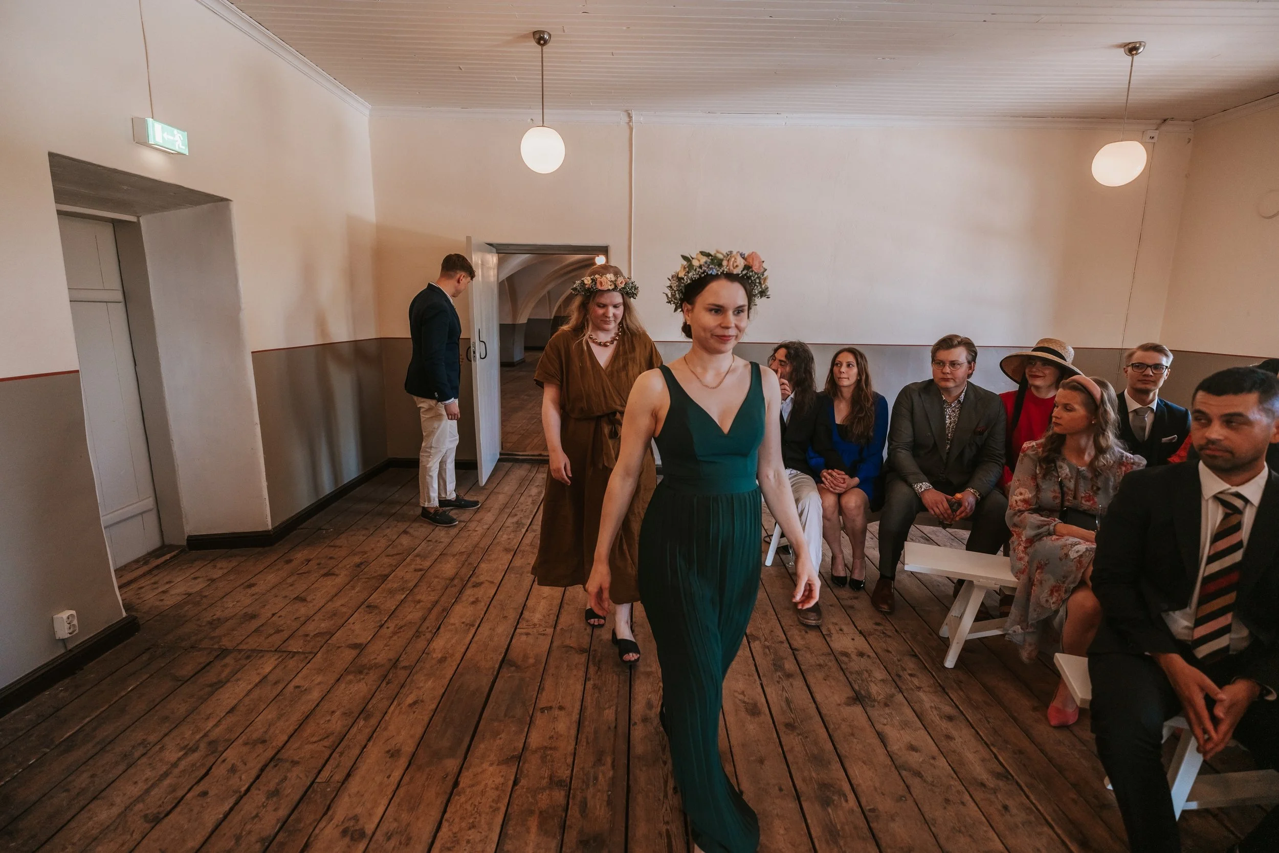 Guests seated in a room watching women in dresses with floral crowns walk by, possibly part of a ceremony.