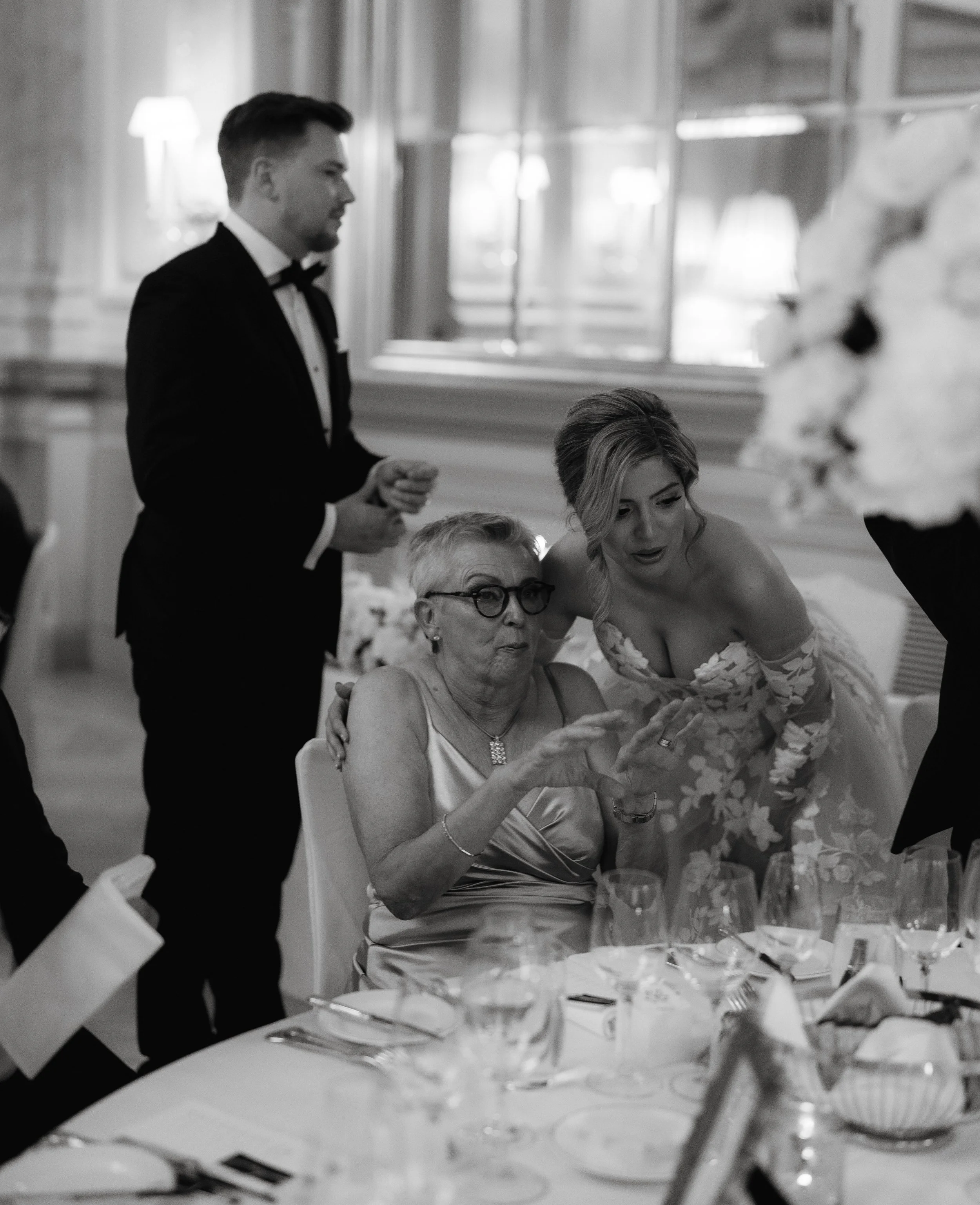 Black and white photo of a formal dining setting with elegantly dressed individuals. A standing man in a tuxedo, a seated woman in a satin dress, and a woman in a floral wedding gown are interacting. The table is set with glassware and cutlery.