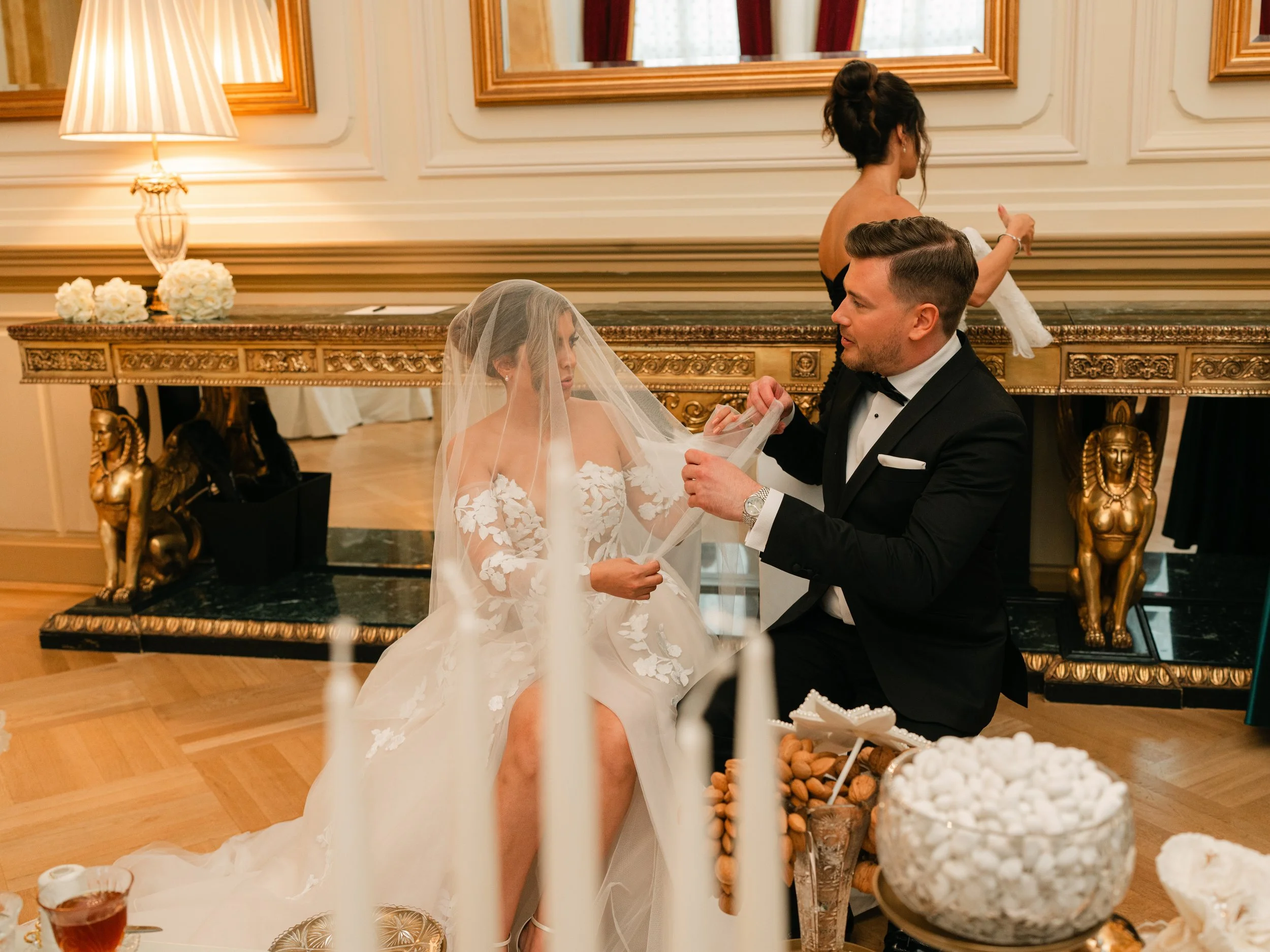 Bride and groom sitting in elegant room with ornate furniture and decorations, woman adjusting bride's veil.