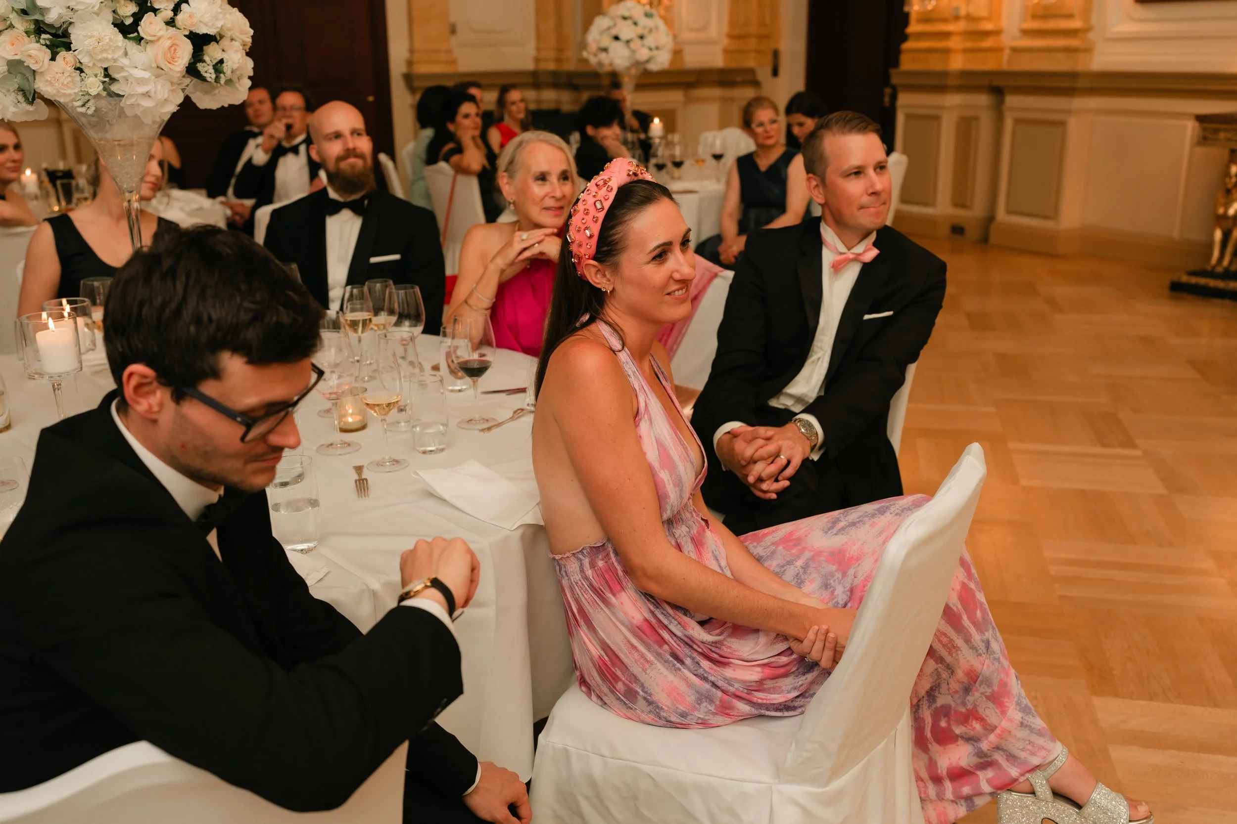 Elegant wedding reception with guests in formal attire seated at tables, floral centerpieces, and glasses of wine.