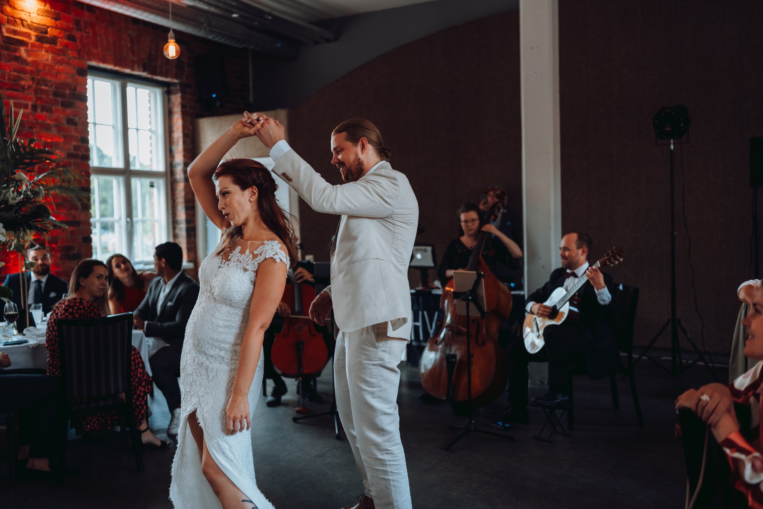 A couple in formal attire dancing at a wedding reception, with a live band playing in the background and guests seated at tables.