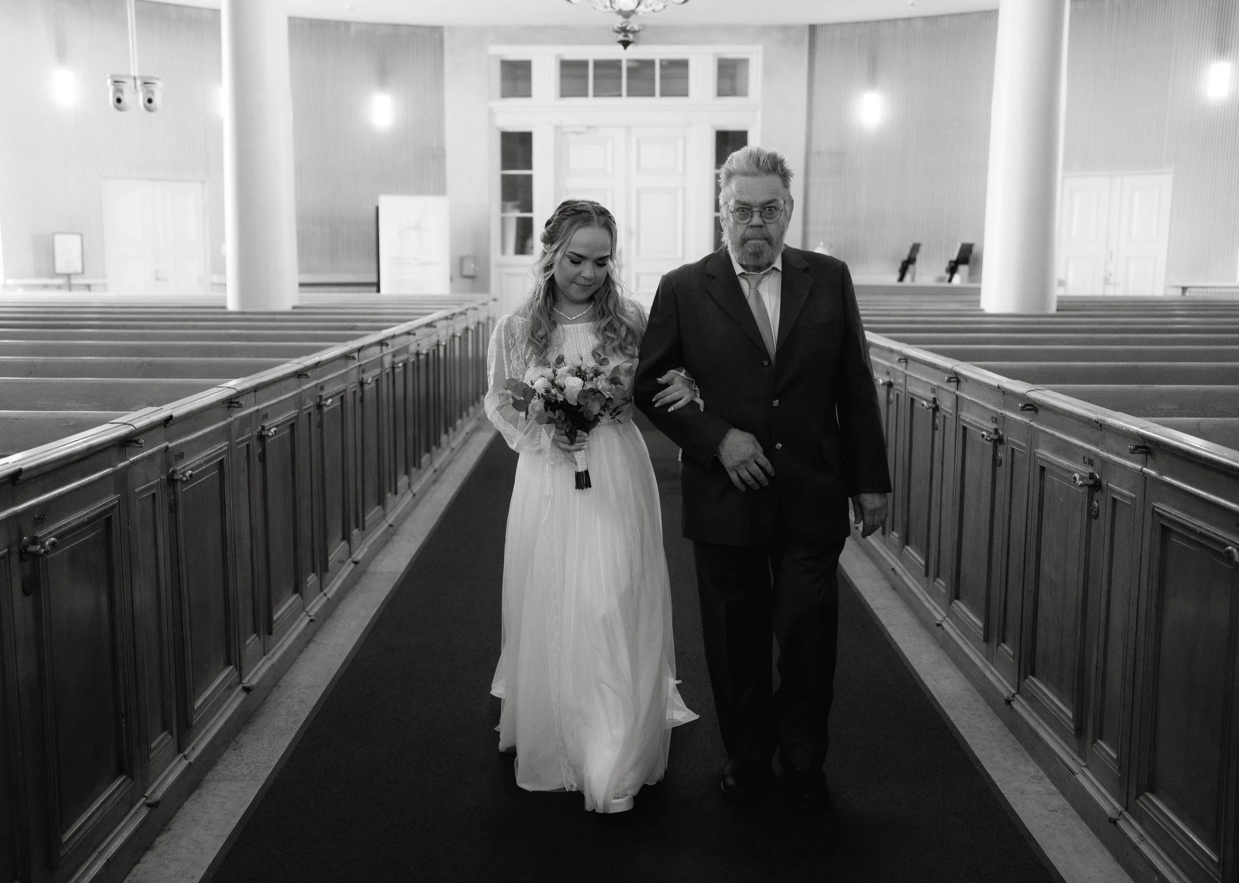 Bride walking down church aisle with older man, holding bouquet, in black and white