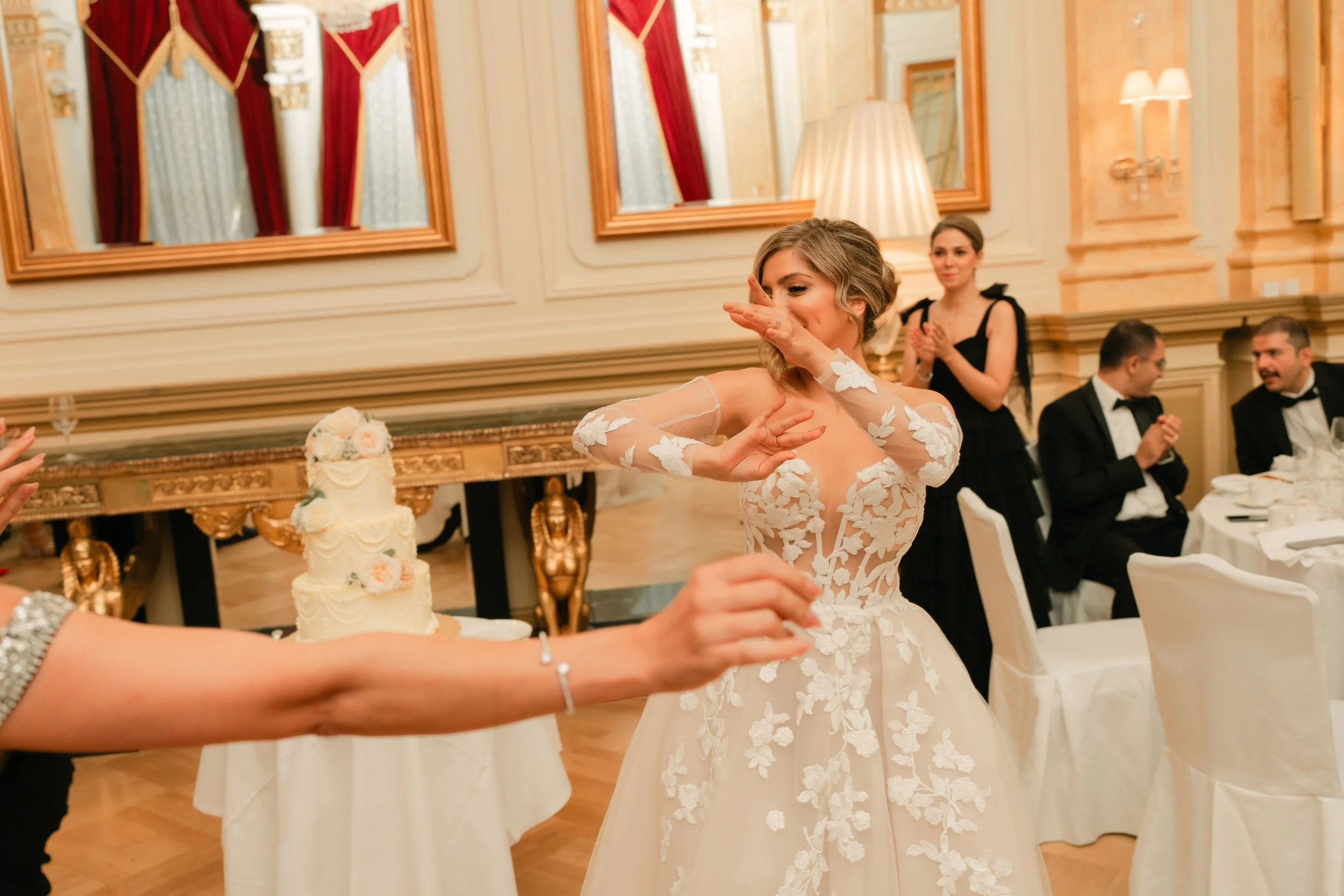 Bride in floral lace gown dancing at a wedding reception, with a tiered cake in the background and guests watching.