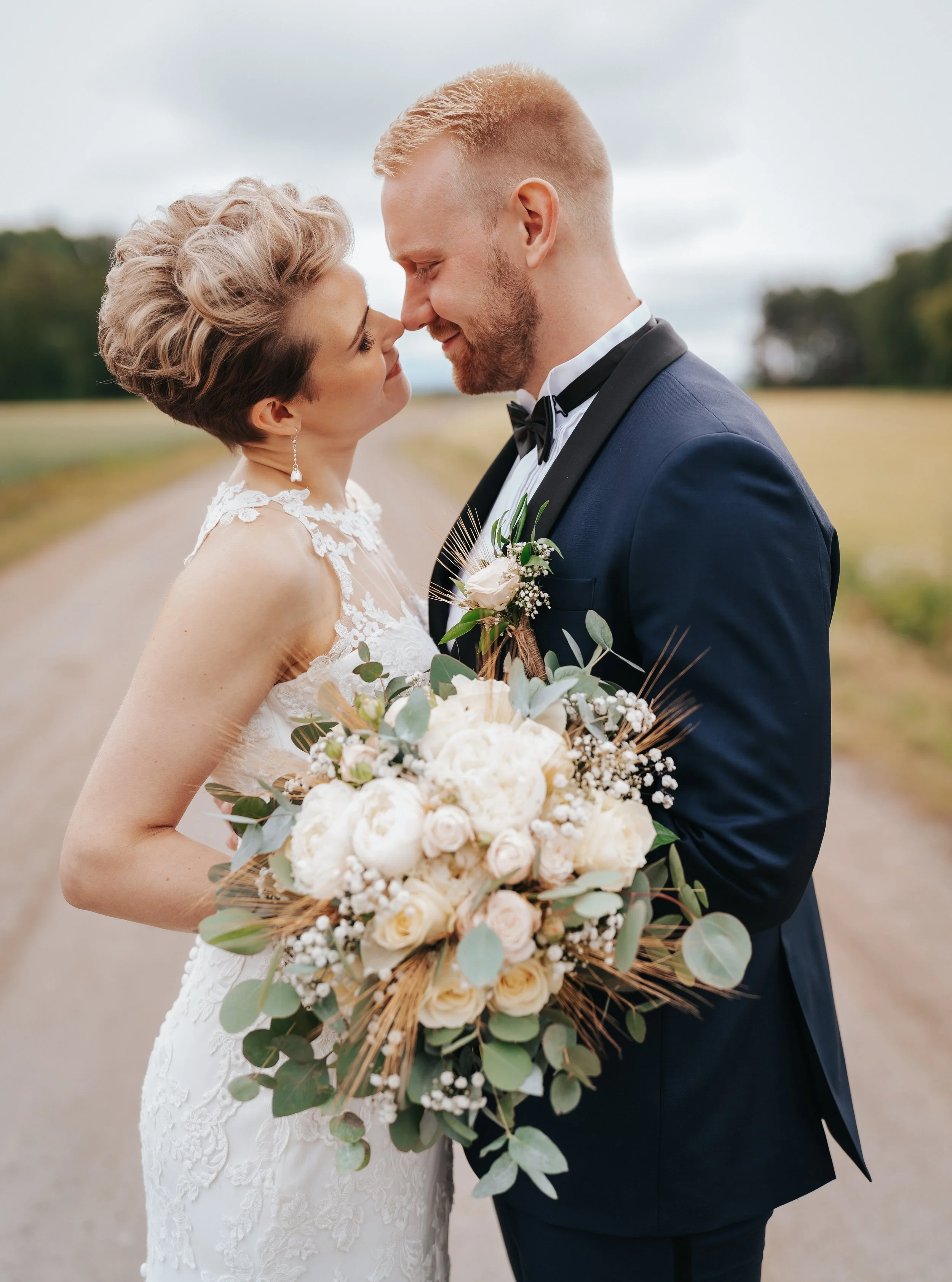 Bride and groom embracing on a rural path, holding a bouquet of white and pink flowers. Bride in a lace dress, groom in a blue suit with a bow tie.