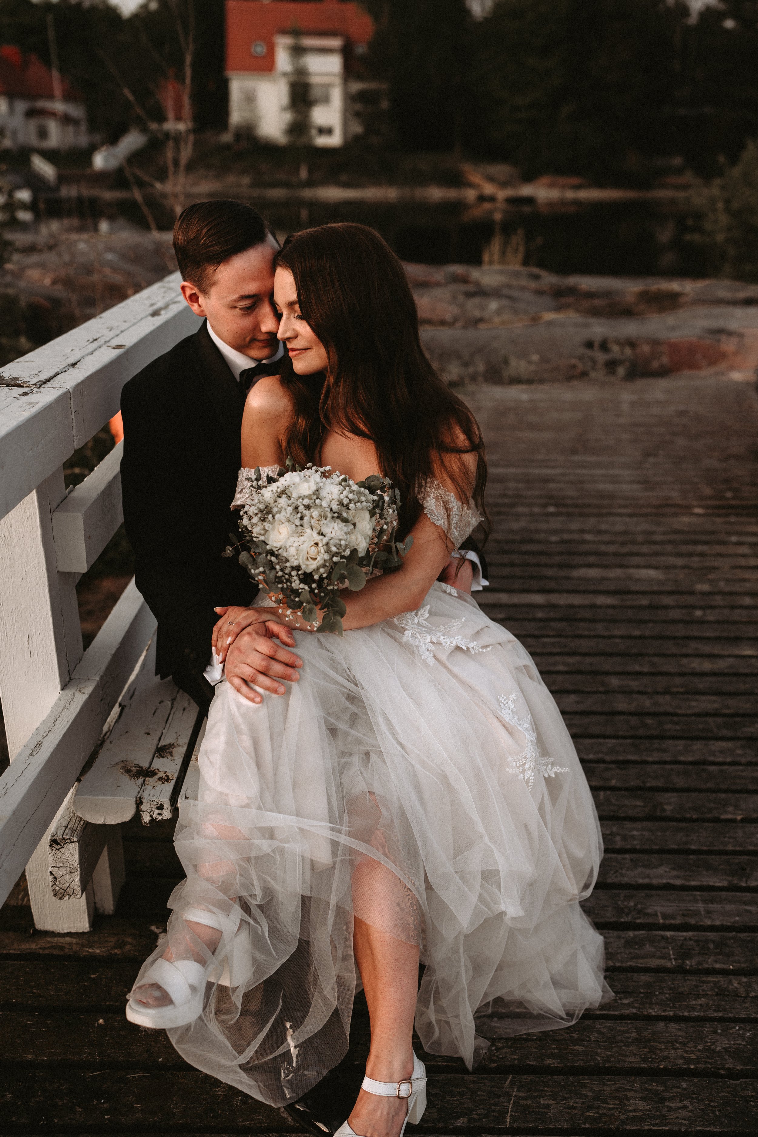 Wedding couple sitting on a bridge, bride in white dress holding a bouquet, groom in dark suit embracing her, romantic outdoor setting.