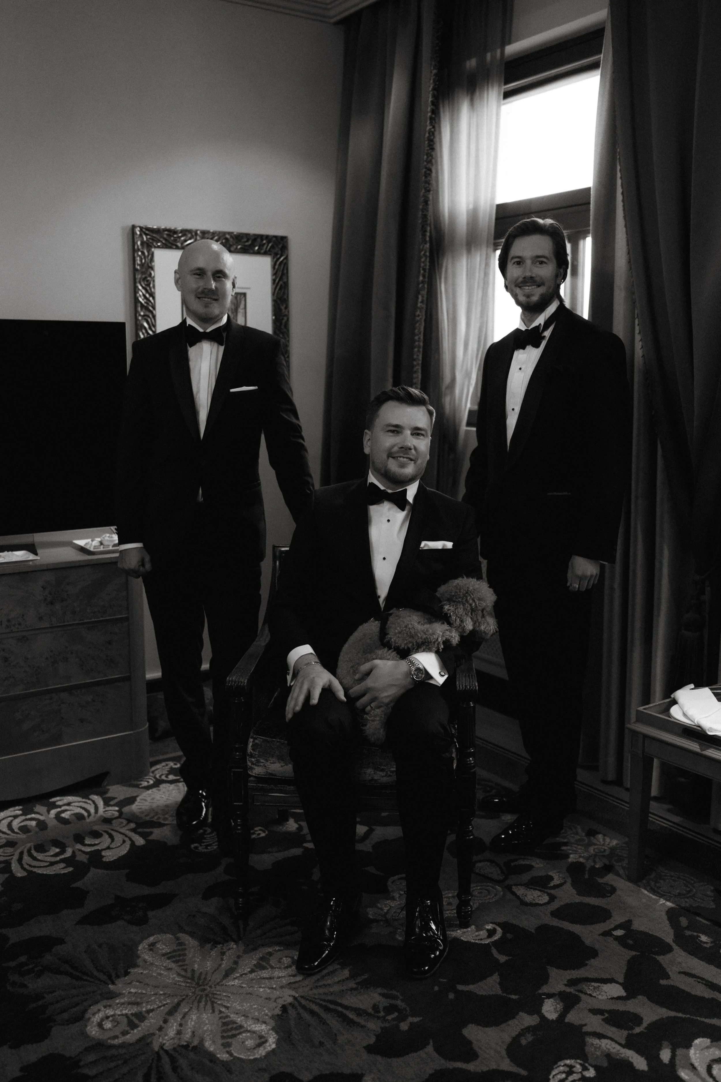 Three men in tuxedos posing indoors; one seated holding a small dog.