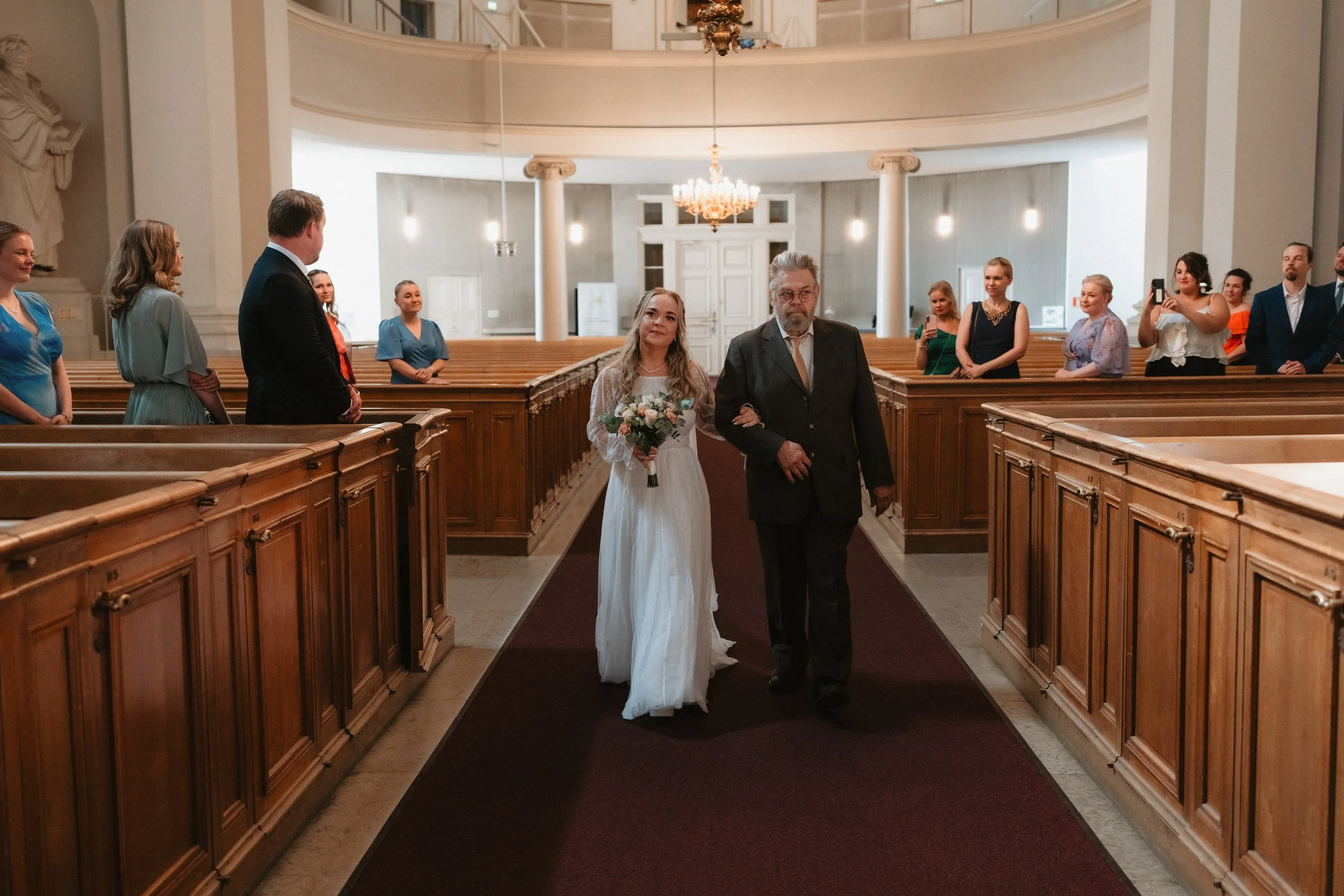 Bride walking down the aisle with an older man in a church setting, surrounded by seated guests.