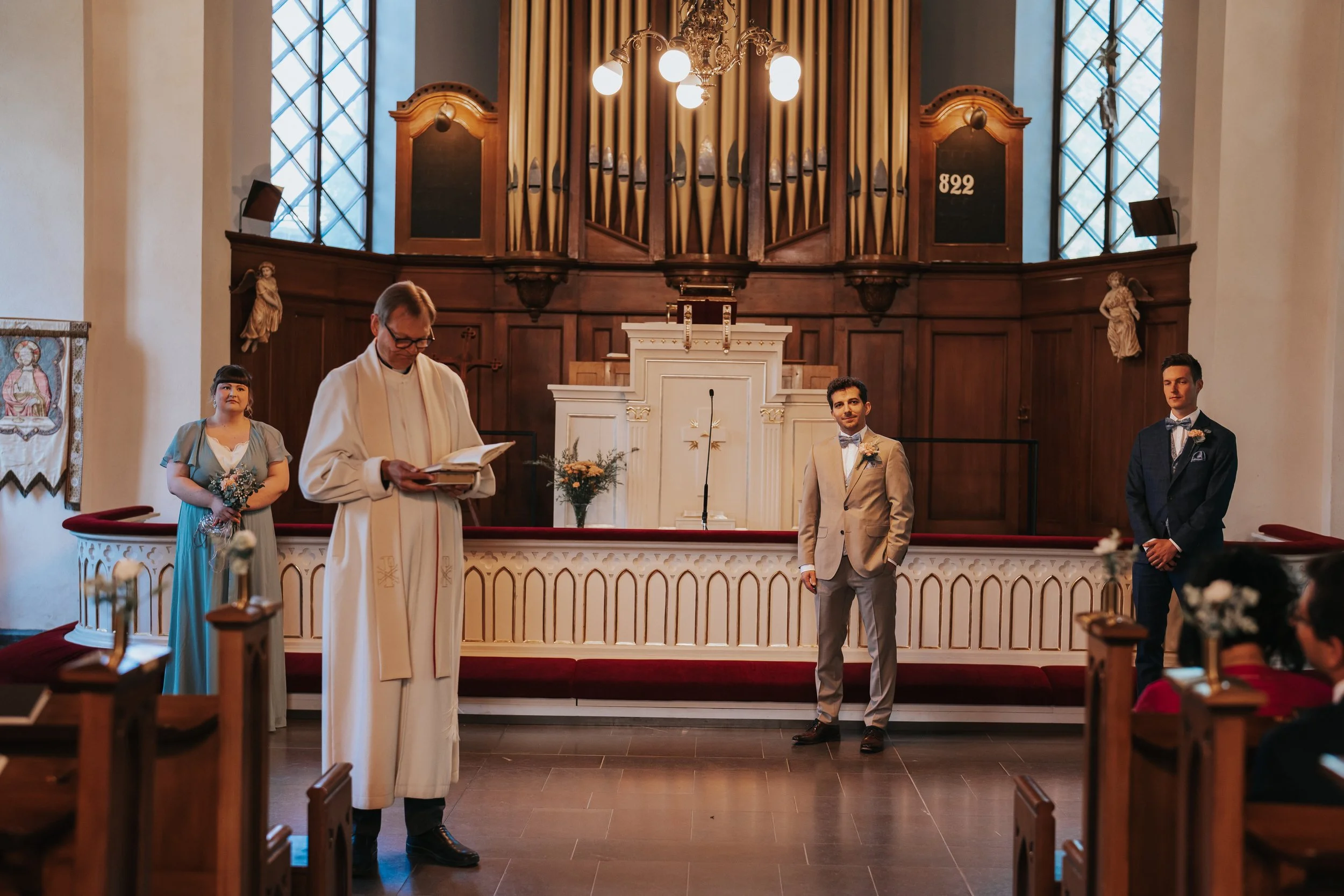 Wedding ceremony in church, with officiant reading, groom in light suit, groomsman in dark suit, and bridesmaid in blue dress.