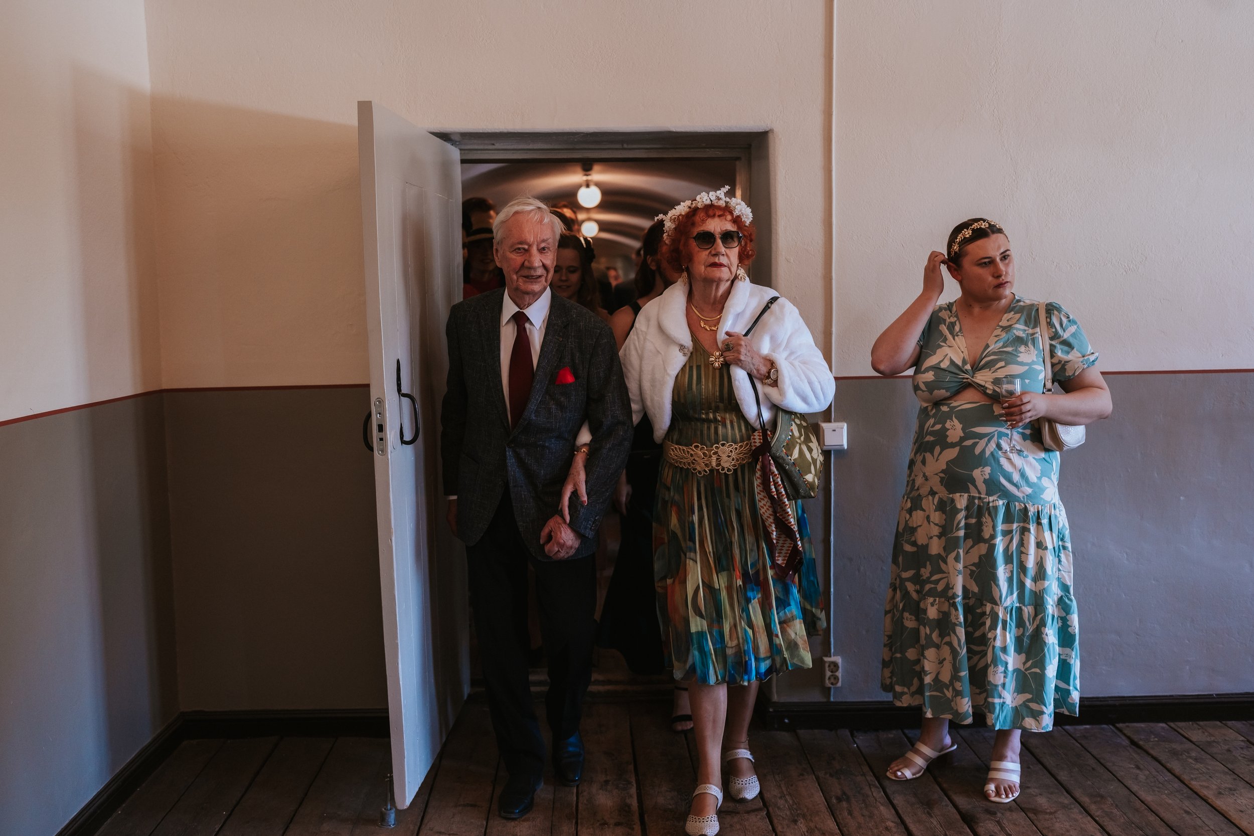 Elderly couple in formal attire walking through a doorway, accompanied by a woman in a floral dress.