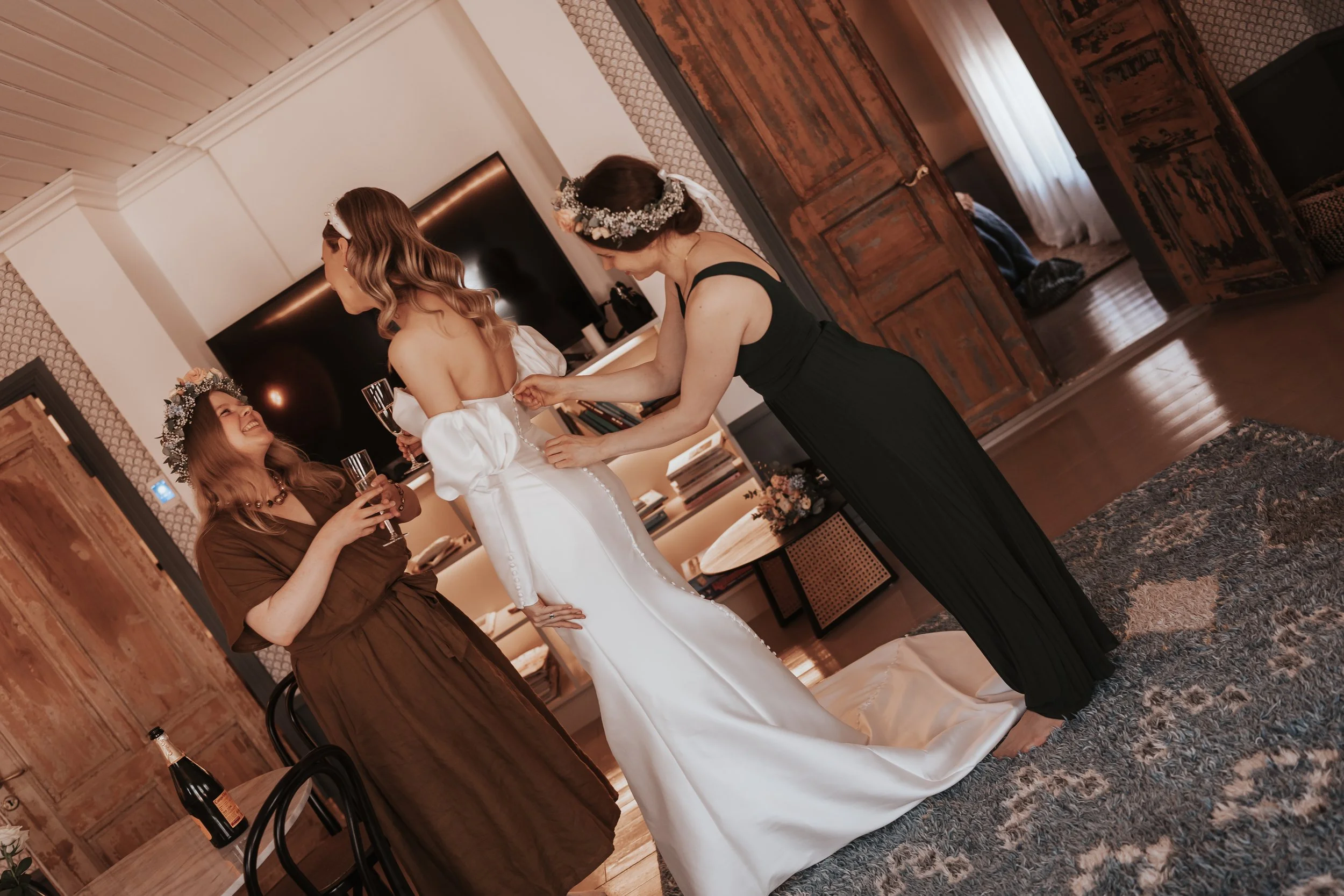 Bridal preparation scene with bride and friends adjusting dress, wearing floral crowns, and holding champagne glasses in a cozy room.