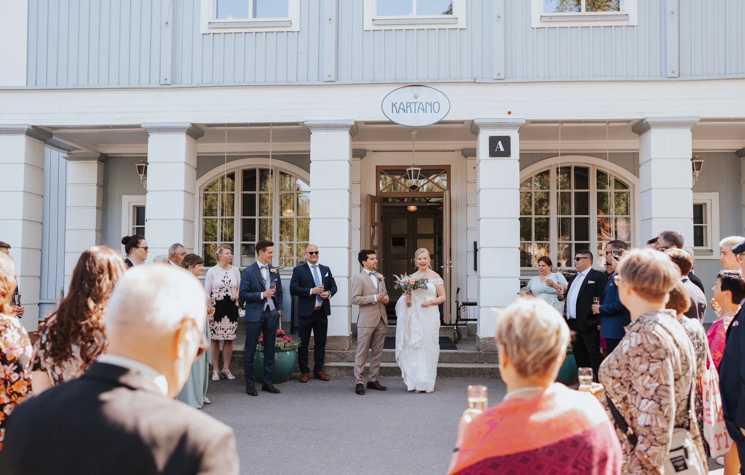Wedding ceremony outside a venue labeled 'Kartano,' with a bride and groom standing together, surrounded by guests in formal attire.