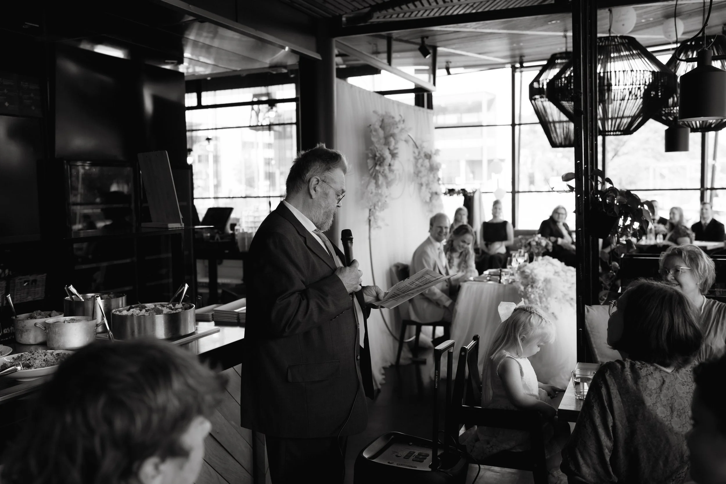 Black and white image of a man in a suit giving a speech with a microphone at a gathering or event. Seated guests, a buffet table, and a decorative backdrop are visible.