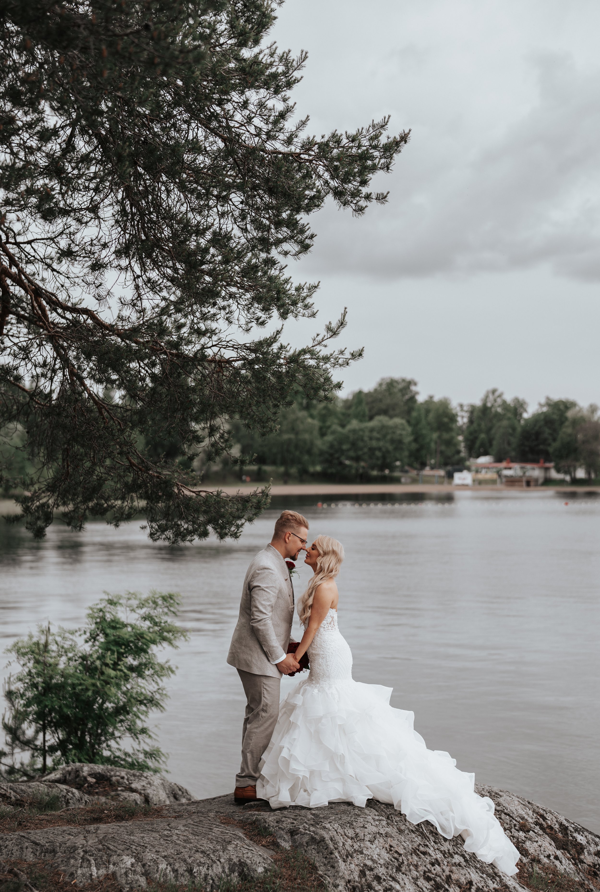 Bride and groom standing on a rock by a lake, surrounded by trees, during an outdoor wedding ceremony.