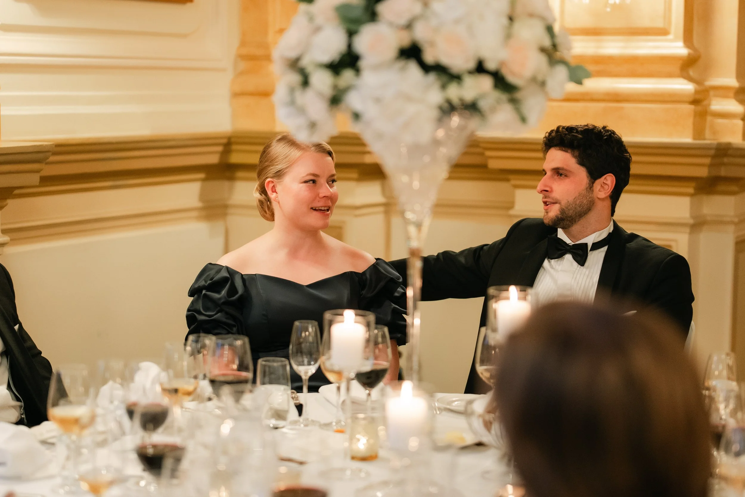 Elegant dinner setting with a man and woman in formal attire seated at a table, surrounded by candlelight and wine glasses, floral centerpiece in foreground.