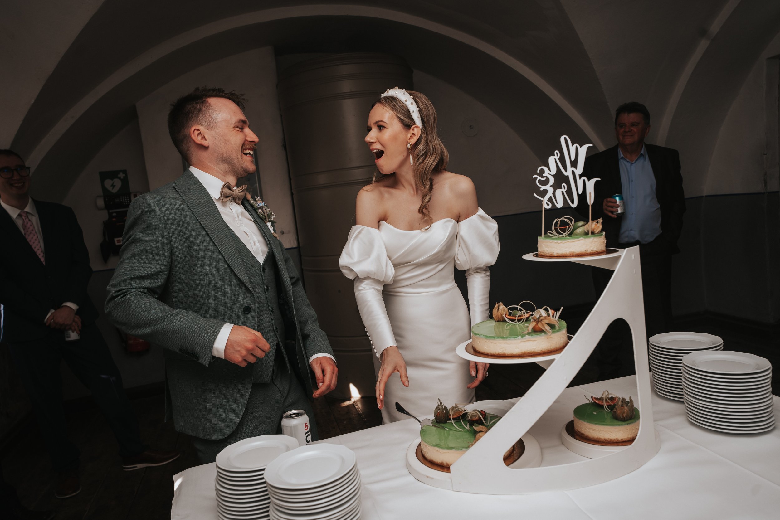 Bride and groom laughing near a tiered wedding cake with green frosting, surrounded by plates and guests.