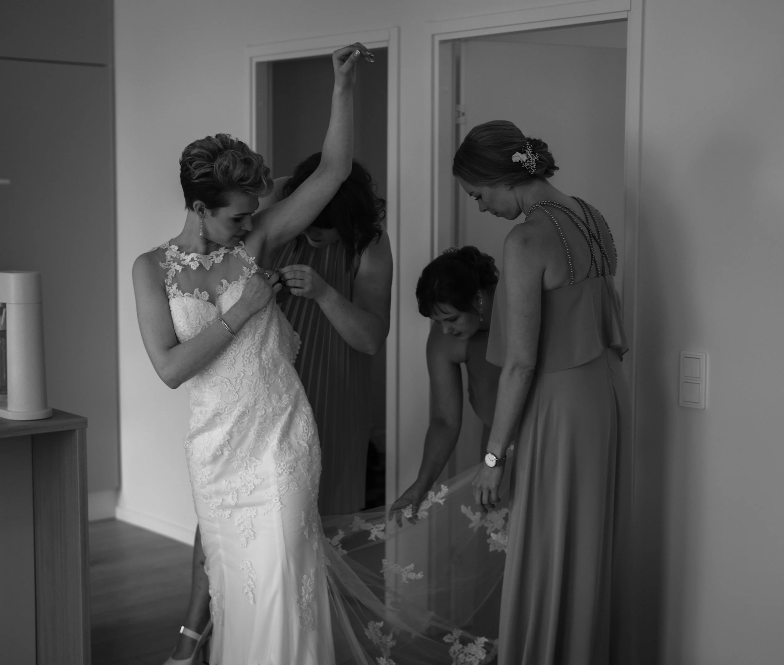 Bride getting ready with bridesmaids adjusting wedding dress in a room.