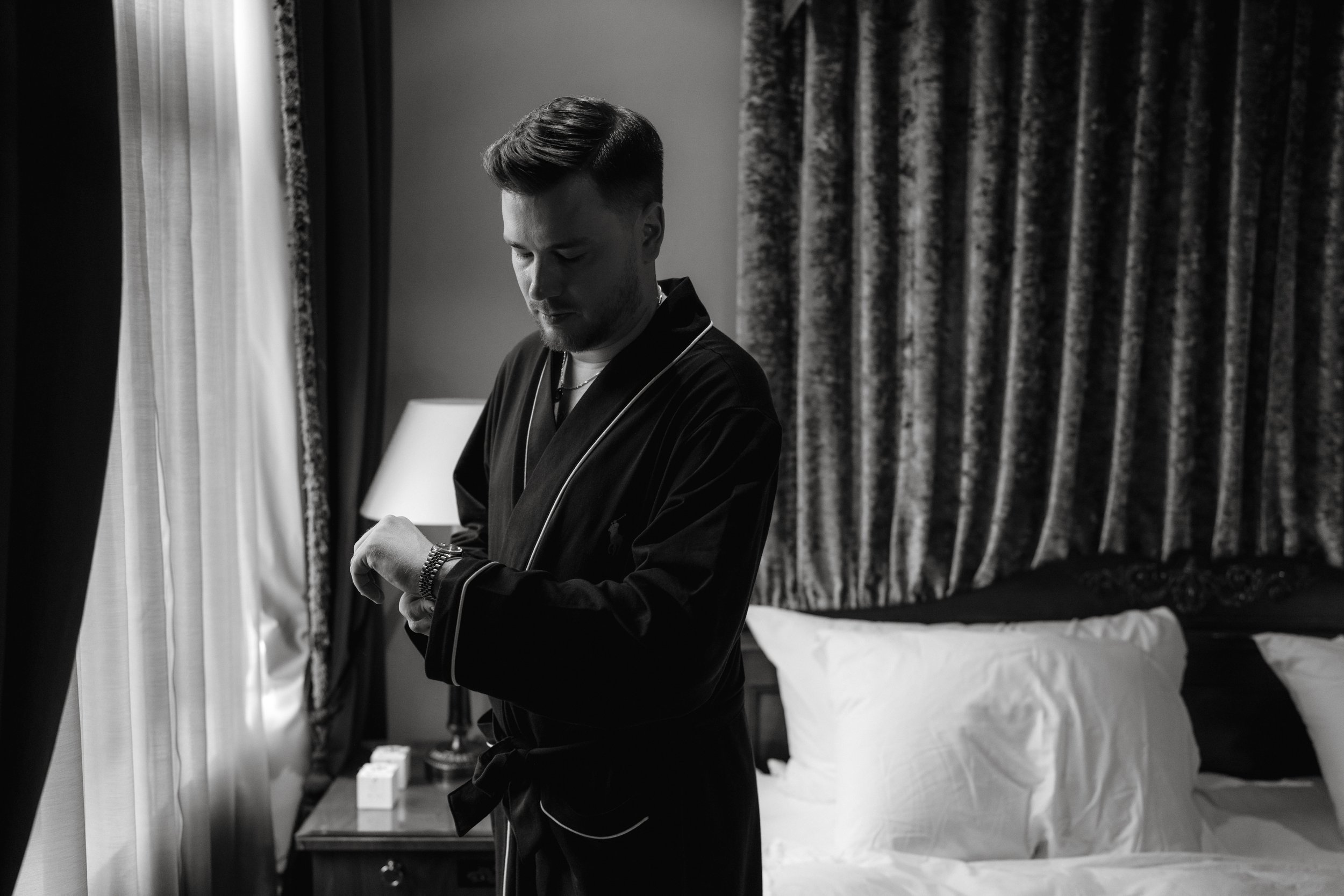 Man in a robe adjusting watch, standing in a bedroom with curtains and a bed in the background.