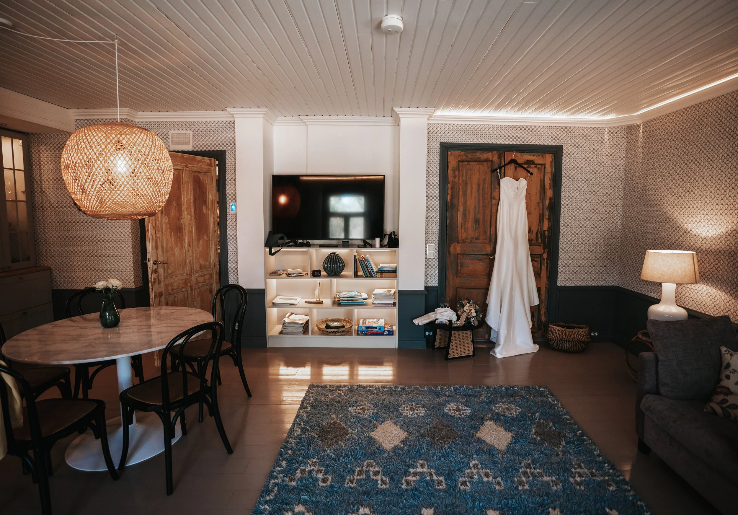 Cozy living room with a hanging wedding dress, modern lighting, a round dining table with chairs, a rug, a wooden door, and a shelf with books and decor items.
