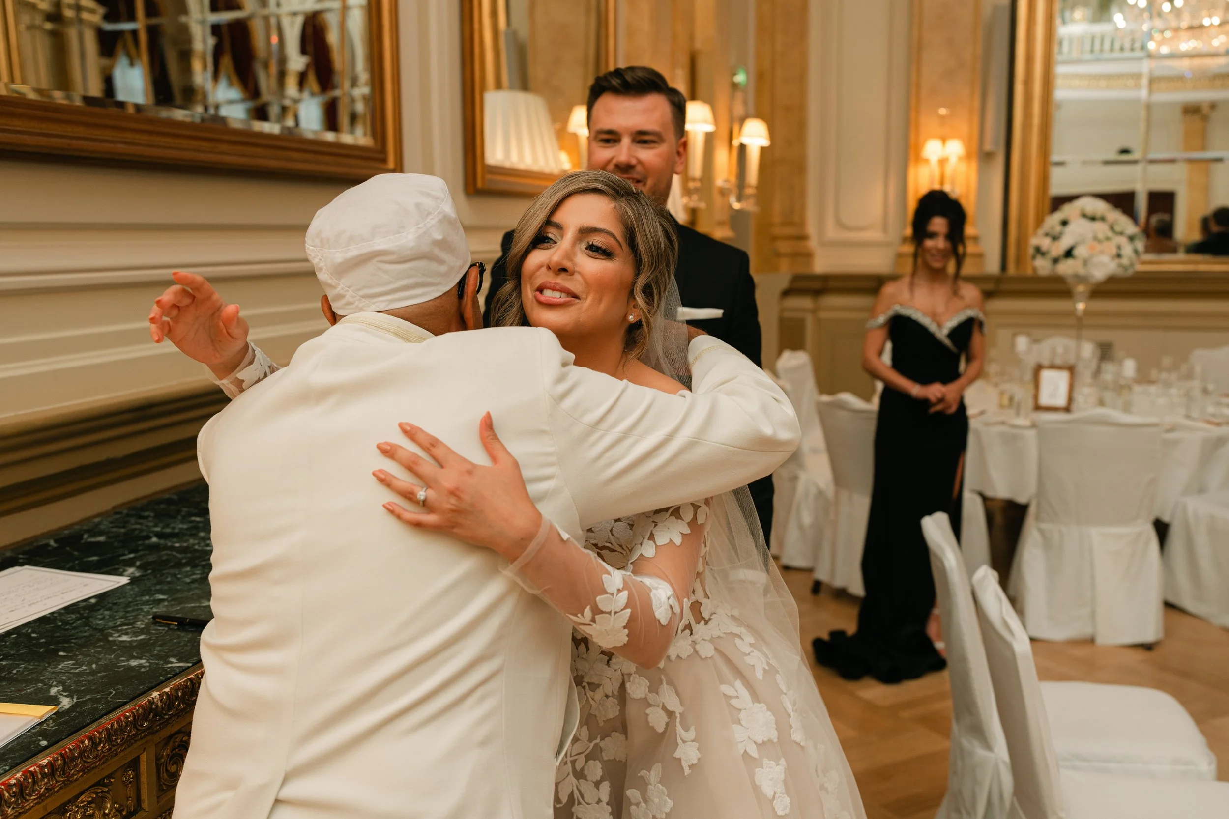 Bride in white dress hugging a person in white attire, with guests in formal wear in an elegant reception venue.