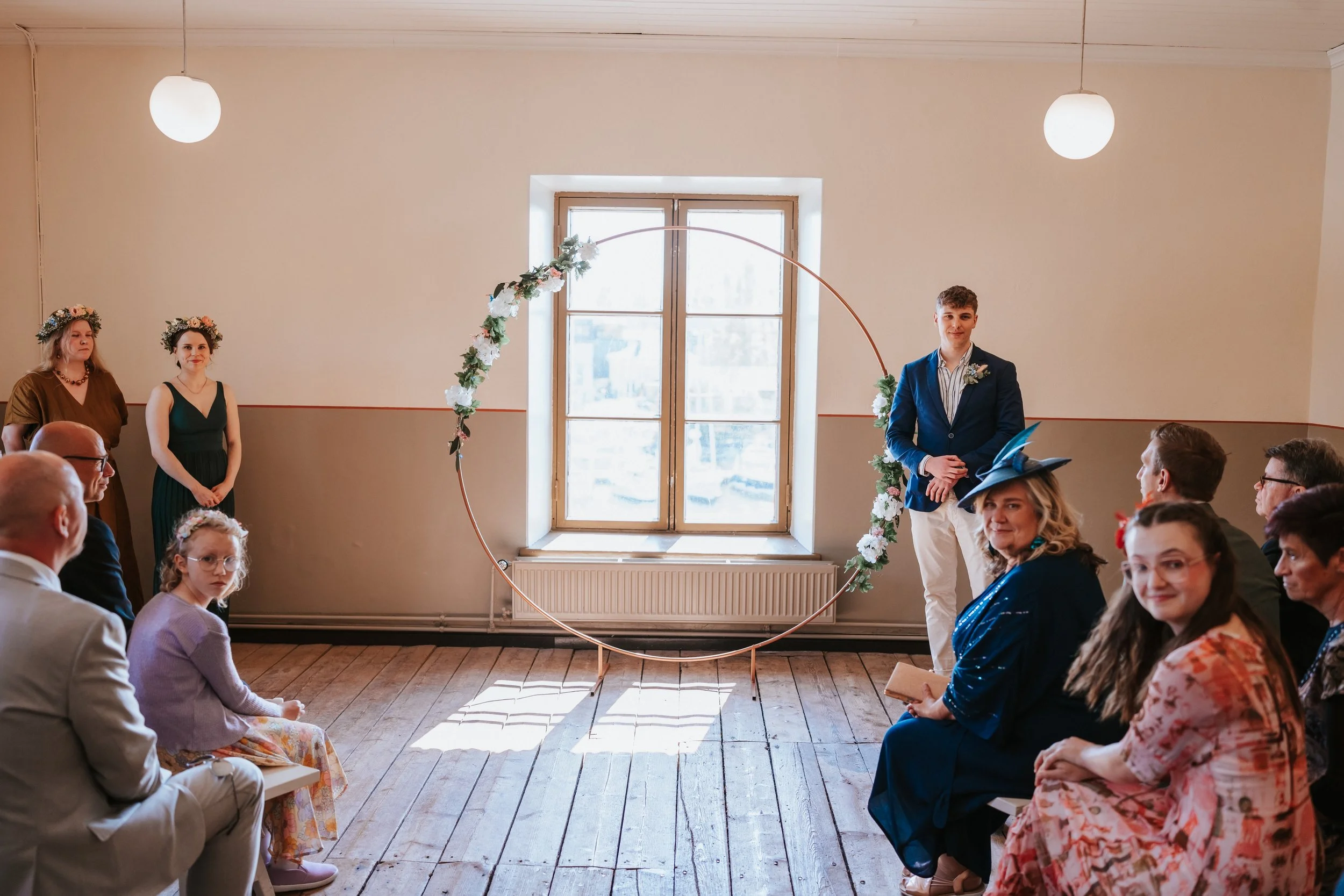 Indoor wedding ceremony with circular floral arch, guests seated, and people standing near window.