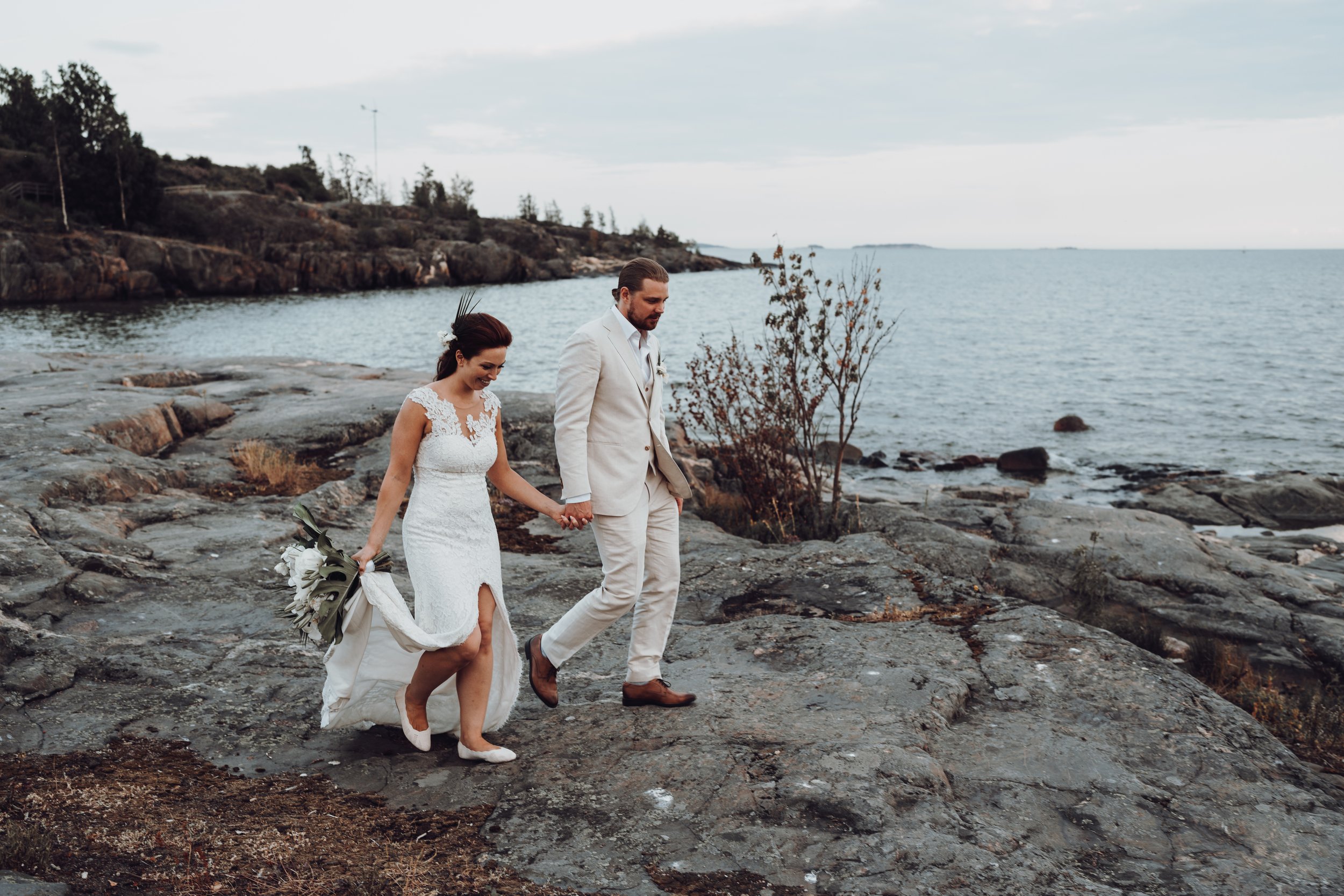 Bride and groom holding hands walking on rocky seashore, ocean in background.