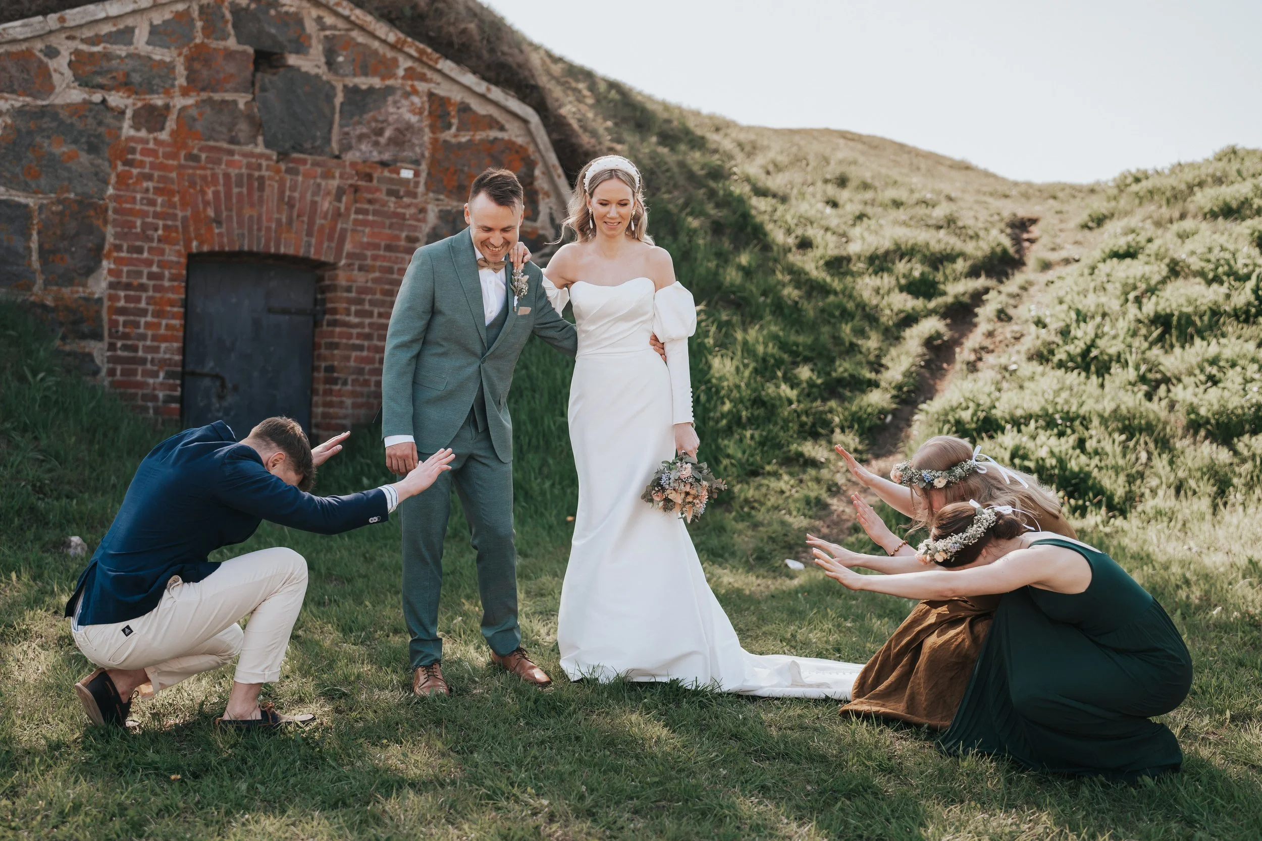 Bride and groom with attendants bowing, outdoors near rustic stone structure.