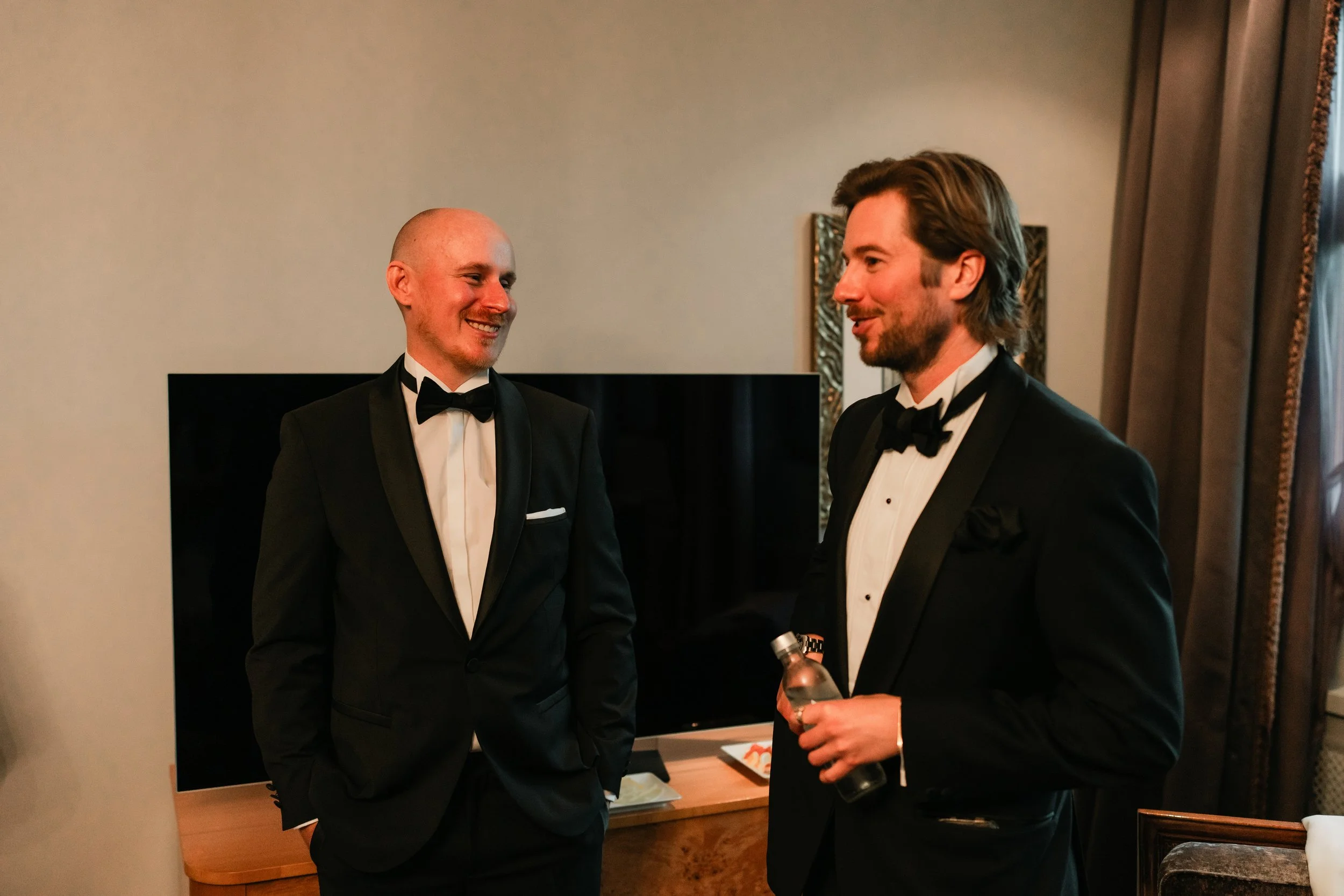 Two men in tuxedos talking and smiling in a stylish room, one holding a bottle, with a framed picture and a TV in the background.