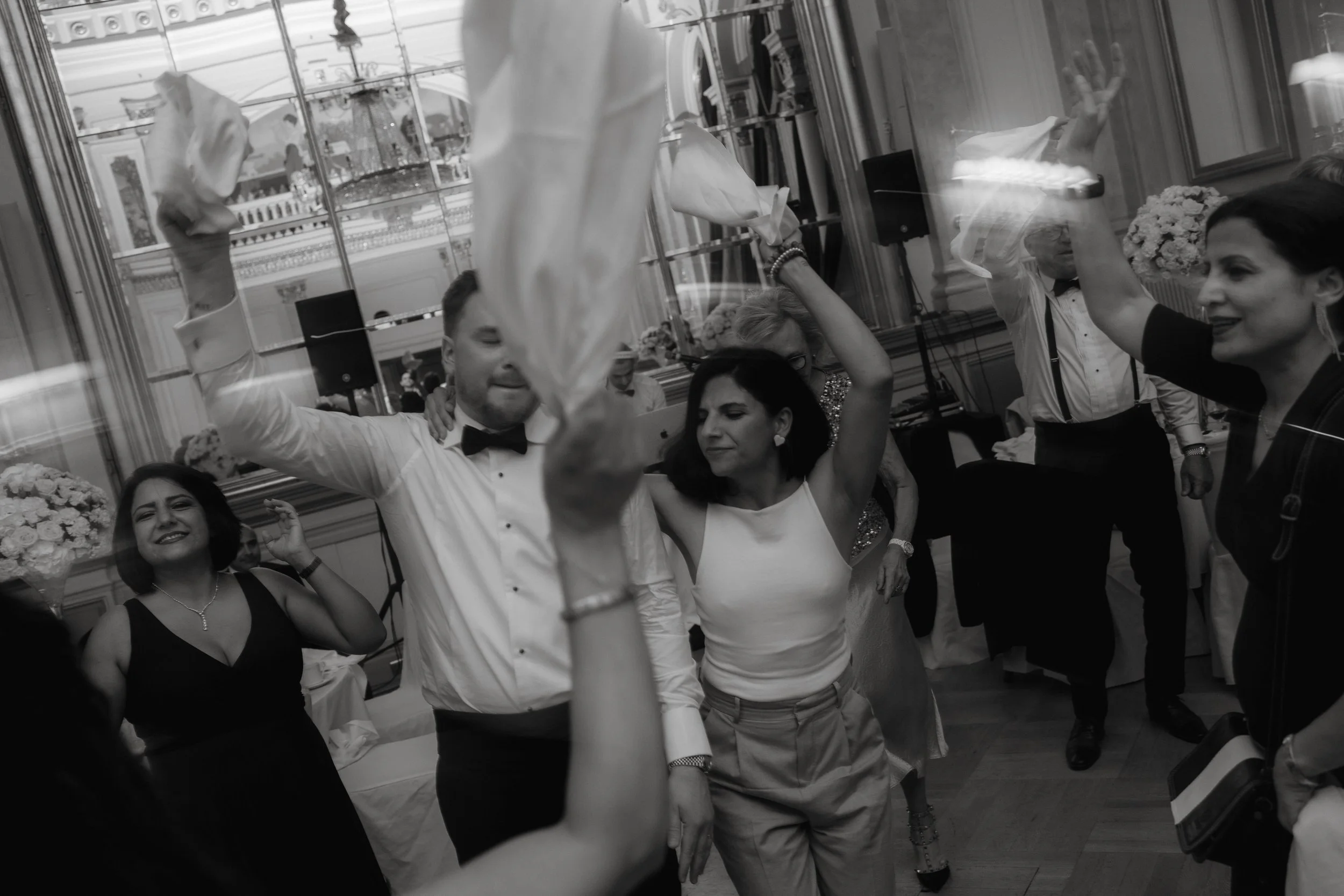 Group of people dancing at a formal event, holding napkins in the air, in a grand hall with floral decorations.