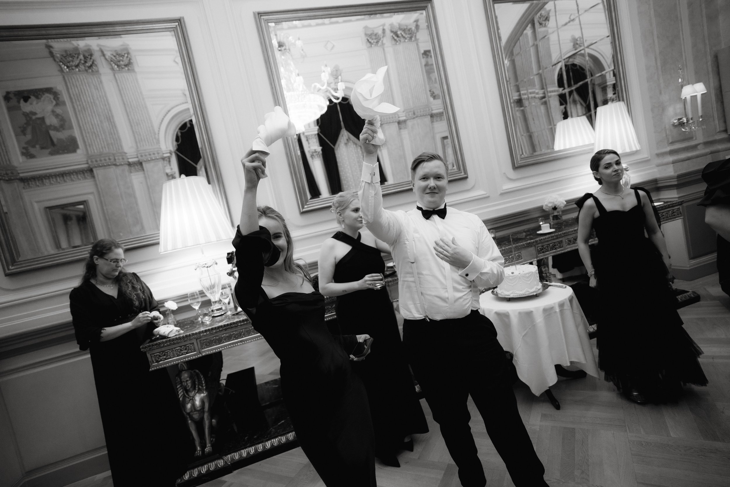 Formal party setting with guests in evening attire, holding napkins in a ballroom with ornate decor and mirrors.