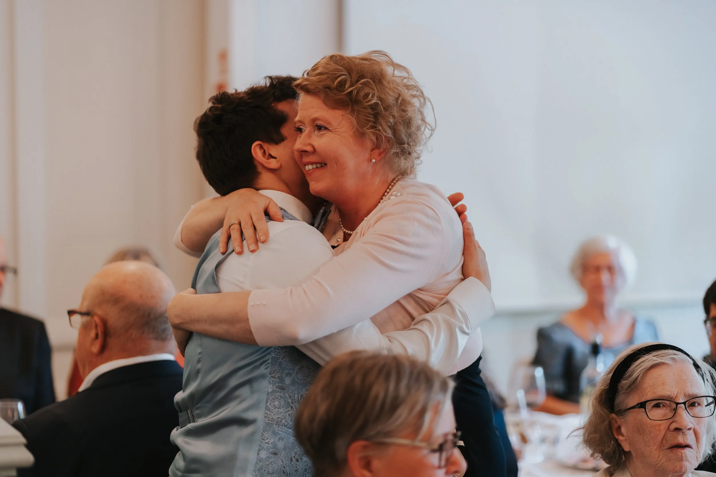 A person in formal attire hugging a woman at an event, surrounded by seated guests.