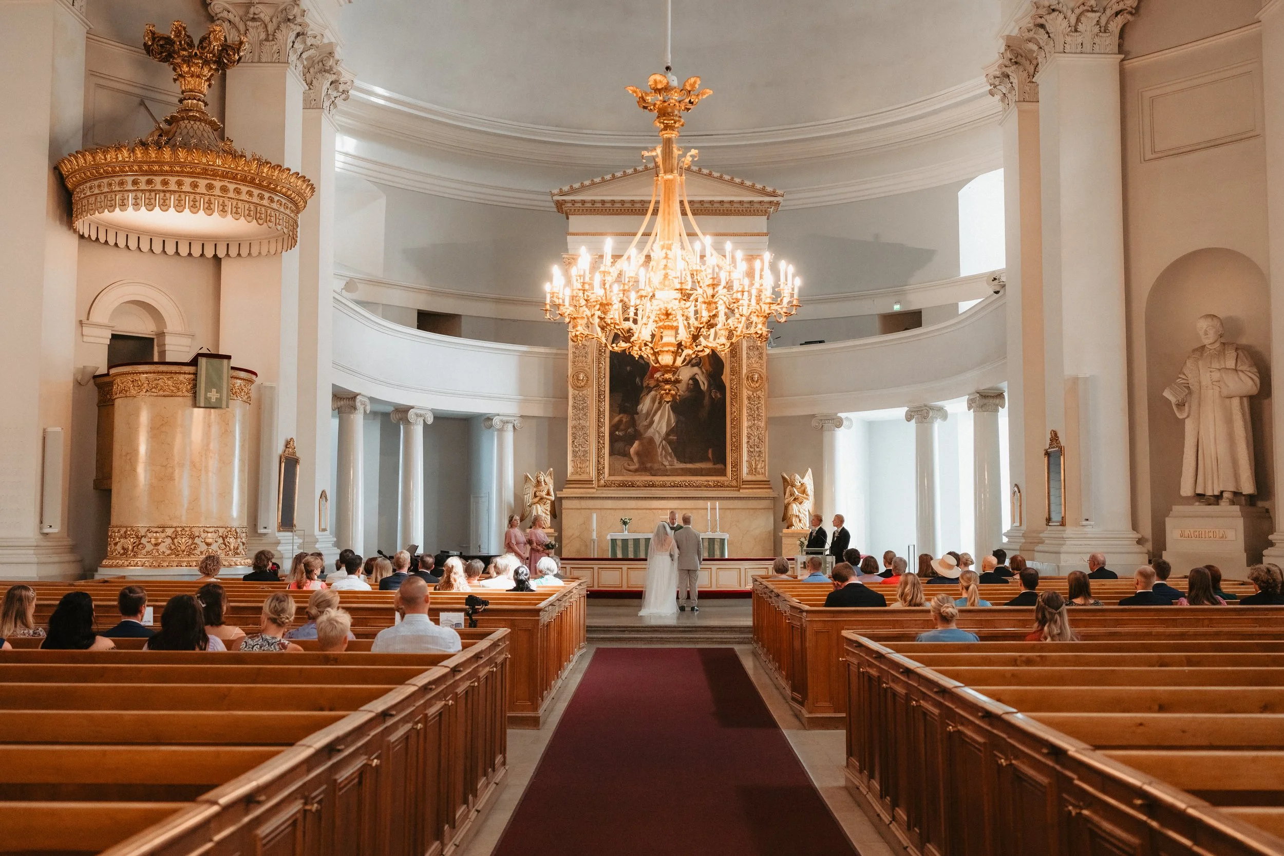 Interior of a church during a wedding ceremony with a large chandelier, pews, and attendees.