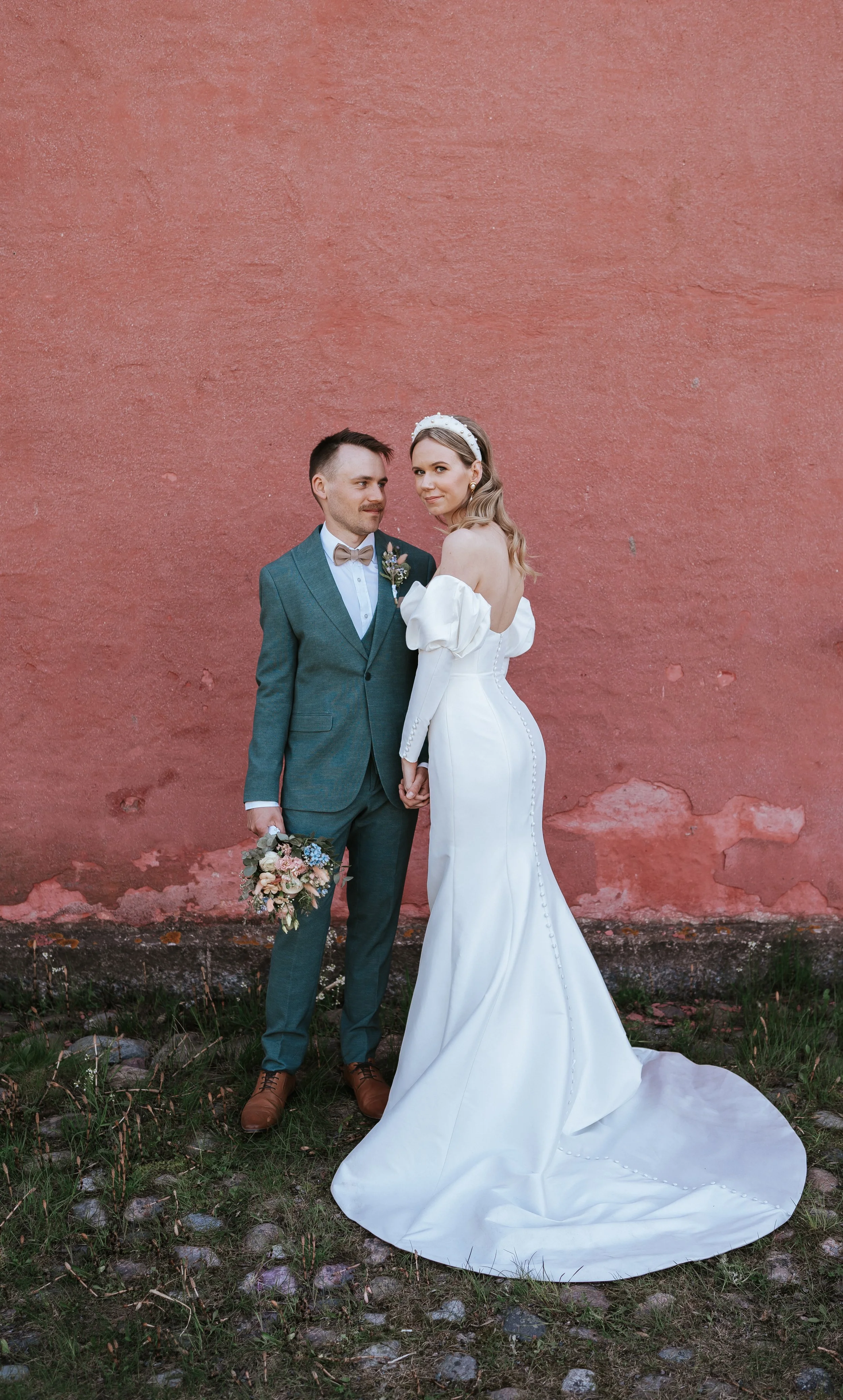 Bride and groom posing against a textured pink wall; bride in a white dress, groom in a blue suit holding a bouquet.